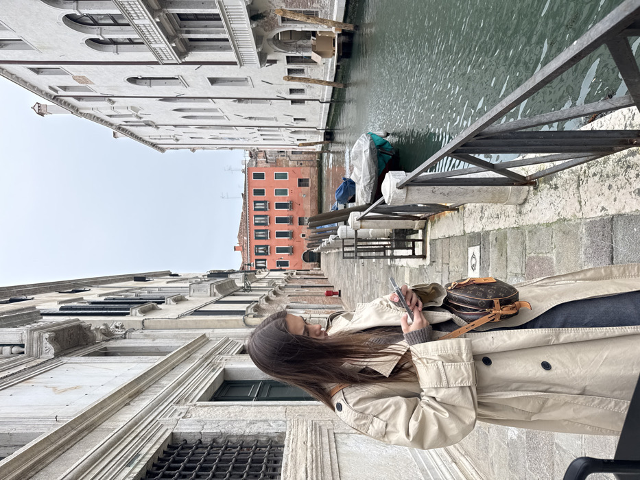A young female stands on a grey stone-paved fondamenta adjacent to a narrow canal in Venice, Italy, under an overcast sky. She has long, dark brown hair and is dressed in a light beige double-breasted trench coat, a dark ribbed sweater, and dark trousers. Her attention is directed downwards as she manipulates a silver-colored smartphone with both hands. A small, brown, monogrammed shoulder bag hangs from her right side.
To her right, a dark metal railing with vertical posts and diagonal cross-members runs along the edge of the canal. The canal contains calm, greenish water, with at least two small boats covered by blue tarpaulins moored to wooden posts near the bank. Buildings characteristic of Venetian architecture line both sides of the canal. On the left, a multi-story building features light-colored stone or stucco, ornate window grates, and decorative moldings. On the right, a multi-story white stucco building displays several arched windows and a small balcony. In the distance, at the end of the canal, a prominent salmon-pink multi-story building with numerous dark-shuttered windows is visible. A red fixture, possibly a fire extinguisher or alarm box, is mounted on the left building's wall further down the fondamenta. No other individuals are present in the visible scene.