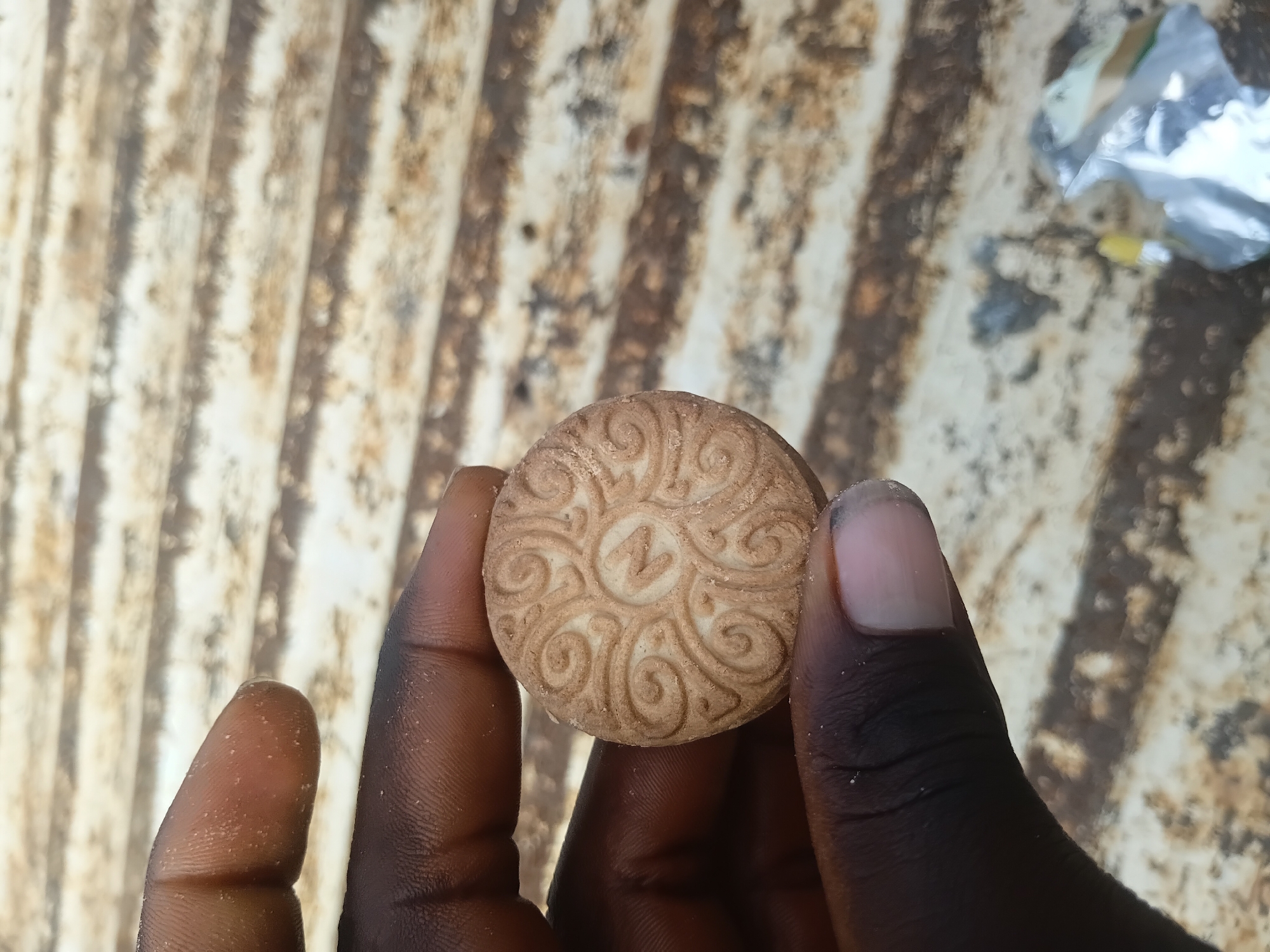 A close-up shot shows a hand holding a round, light brown cookie with a detailed embossed pattern. The pattern features swirling lines and a central design that resembles the letter "N" enclosed in a circle. The cookie has a slightly crumbly texture. The background is out of focus, appearing to be a textured surface with horizontal lines, possibly wood or metal. A crumpled piece of foil and a small yellow object are visible in the lower right corner of the image.