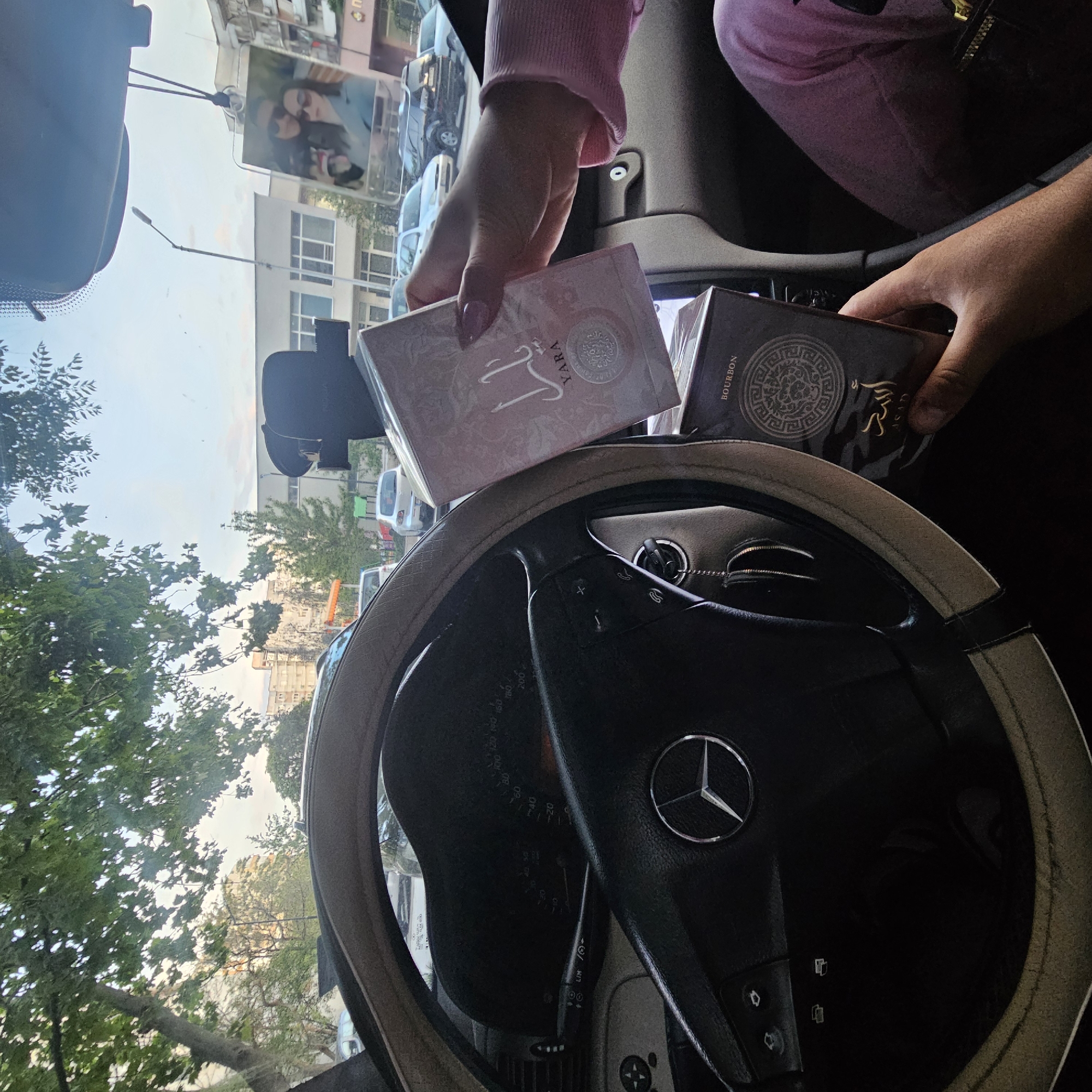 Here's a forensic description of the image:
**Scene:** Interior of a car, likely a Mercedes-Benz (based on the steering wheel logo) on a street. Possibly Plovdiv, Bulgaria, due to the Cyrillic text on a nearby storefront visible through the windshield.
**Subjects:**
* Two hands are visible holding two packaged perfumes. One box is light pink with the word "YARA" on it. The other box is dark brown/black with the word "BOURBON" on it.
**Objects:**
* Mercedes-Benz steering wheel with a light-colored cover.
* Car dashboard.
* Two perfume boxes as previously described.
* Windshield mounted device, possibly a phone holder or navigation system.
* Windshield, revealing a street scene outside.
**Environment:**
* Urban environment with buildings and parked cars visible through the windshield.
* Trees and vegetation visible through the windshield.
* Signs of a store written in Cyrillic, likely Bulgarian.
**Actions/Interactions:**
* A person is holding two perfume boxes over the steering wheel.
**Additional details:**
* The driver is holding perfumes that is called YARA and Bourbon.