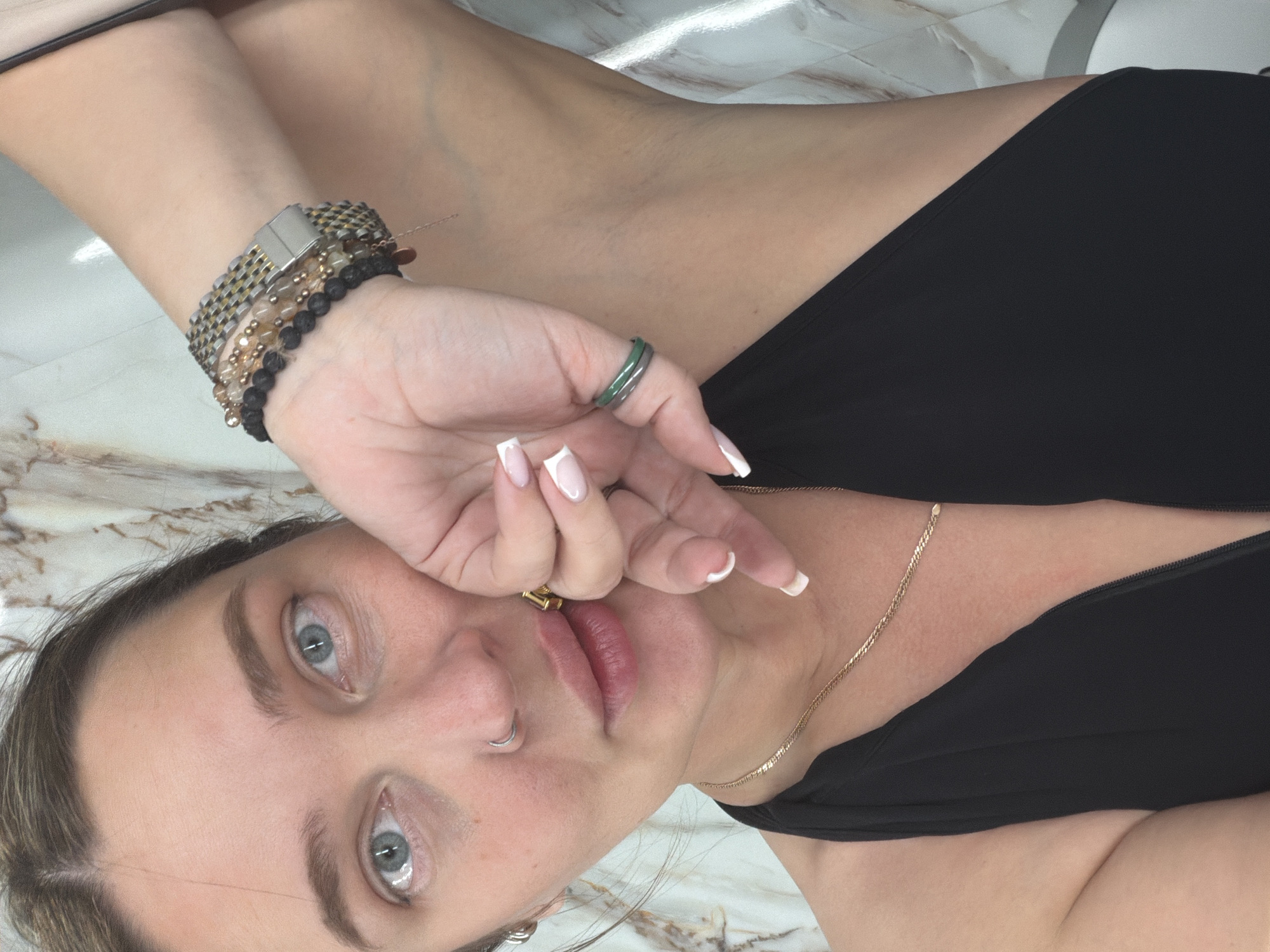 A close-up, angled view of a woman with light skin and light blue eyes. She is wearing a black top. Her left hand is raised to her face, adorned with a watch and several beaded bracelets. A green ring is on her left ring finger. She has a gold chain necklace and a nose ring. Her nails are painted with a French manicure. The background appears to be a light-colored patterned surface. The location is not discernible from the image. The image does not depict a location in Moscow, Russia.