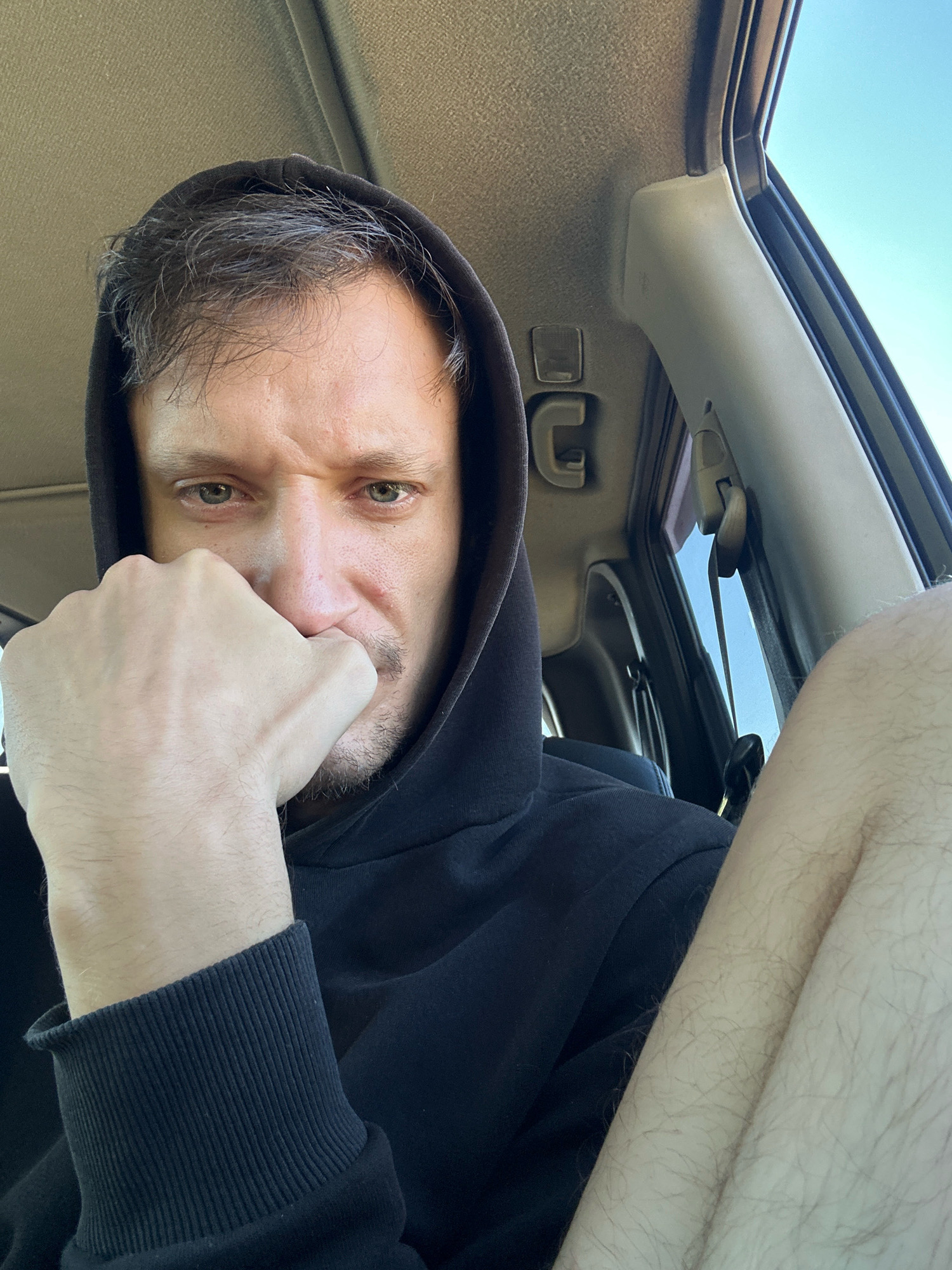 A man wearing a black hooded sweatshirt is inside a vehicle, his left hand resting on his face near his mouth. His eyes are green and he has a light mustache. The interior of the car is visible, including the ceiling and a portion of the passenger seat. The man's right leg is also visible, covered in light hair. The scene appears to be outdoors on a sunny day, with the sky visible through the car window. The location is not specified as Makiv, Ukraine.