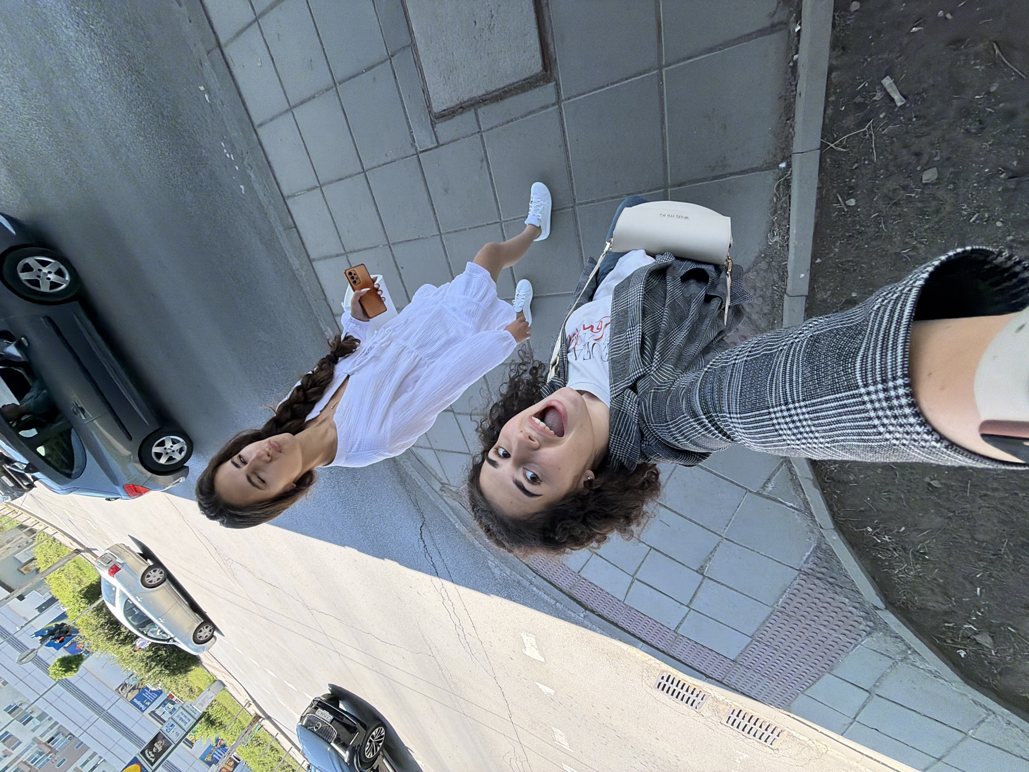 The image depicts two young women on a city sidewalk in Sofia, Bulgaria. The woman in the foreground, with curly dark hair and wearing a plaid jacket over a white t-shirt, is taking a selfie with an open-mouthed expression. She is carrying a small, light-colored handbag. Behind her, the second woman, with long dark braided hair, wears a white dress and white sneakers, and is holding a mobile phone in her right hand. She is looking to her left with her lips pursed.
The background shows a city street with vehicles, including a silver sedan and a dark-colored sedan. A grey hatchback is parked on the street to the right of the women. The sidewalk is paved with square tiles, and there are metal grates and tactile paving visible. The overall environment suggests a bright, sunny day in an urban setting.