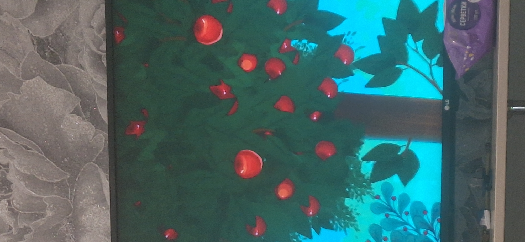 The image shows a close-up of a television screen displaying a cartoon of a tree with red apples and green leaves against a bright turquoise background. The tree trunk is brown and appears to be in the center of the frame. Some of the apples are round and red, while others are depicted as red crescent shapes. There are also red leaf-like shapes scattered among the apples. The bottom of the screen shows the LG logo and part of a shelf with a purple package of wet wipes with Russian text, specifically "СЕРБЕТКИ" which translates to "wipes". Above the television screen, a textured wallpaper with a floral pattern is visible. The overall scene is a still image from a television display. No specific location in Bila Tserkva, Ukraine can be determined from the visual information provided.
