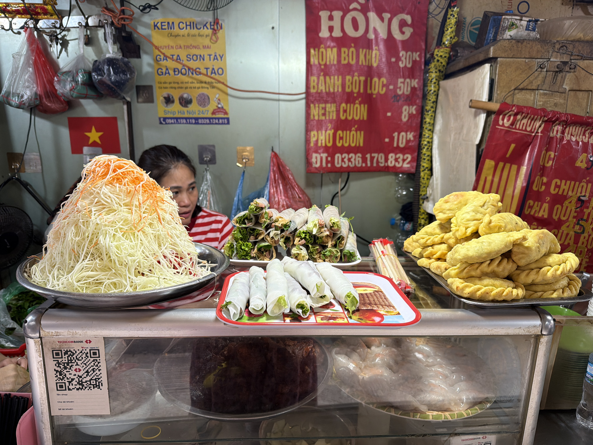 The image shows a street food vendor in Hà Nội, Vietnam. A woman in a red and white striped shirt stands behind a display case filled with Vietnamese dishes, including a large pile of shredded papaya salad, fresh spring rolls (gỏi cuốn), and fried dumplings (bánh gối). A sign above advertises "KEM CHICKEN" and various types of chicken, along with phone numbers for ordering. Another sign lists prices for items like "NỘM BÒ KHÔ" (dried beef salad), "BÁNH BỘT LỌC" (tapioca dumplings), "NEM CUỐN" (fresh spring rolls), and "PHỞ CUỐN" (rolled pho). The Vietnamese flag is visible in the background. The display case has a Techcombank QR code for mobile payments.