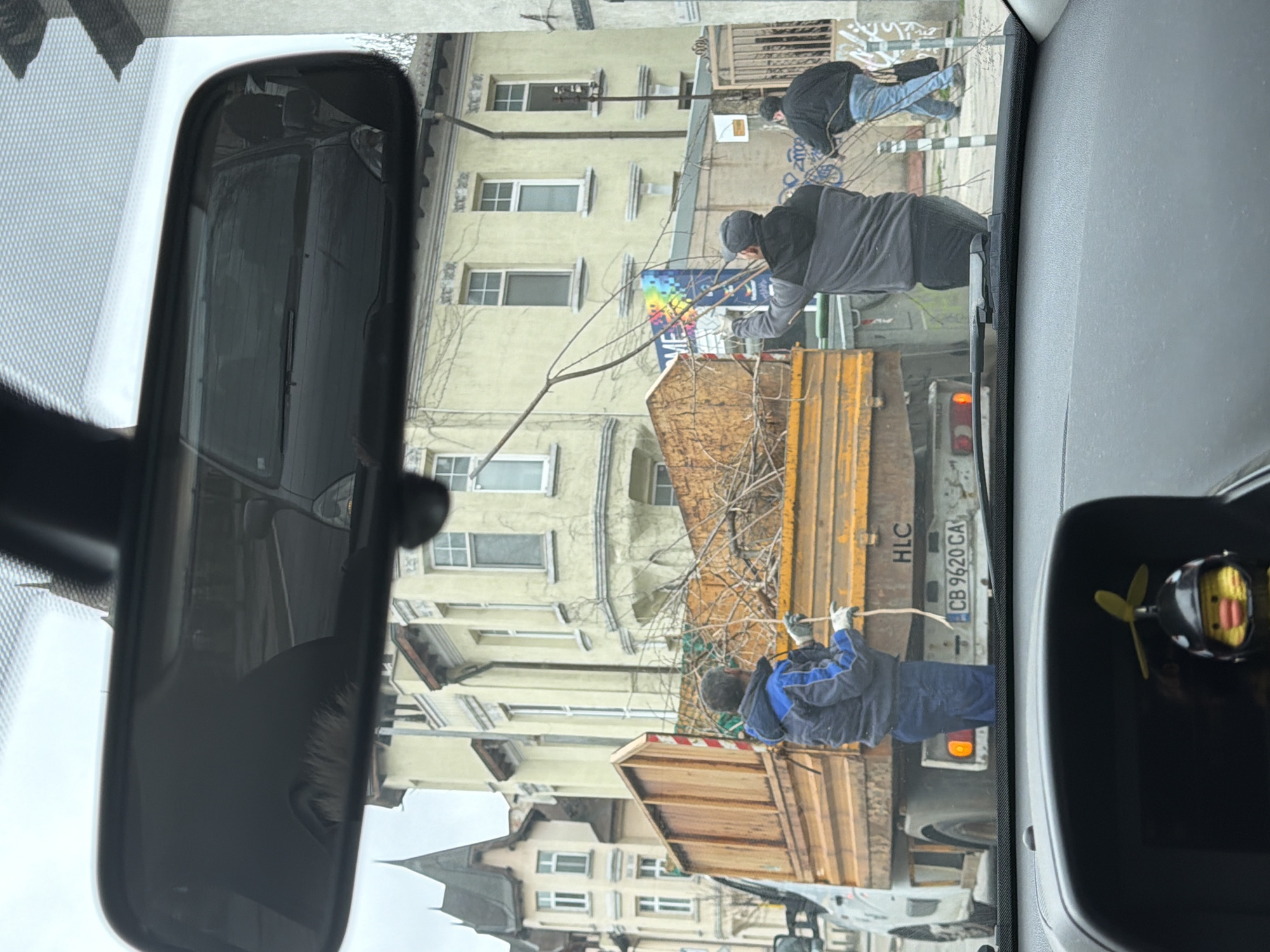The image, taken from the interior of a vehicle, captures a scene outside featuring a yellow flatbed truck bearing the license plate "CB 9620 CA". The truck bed is filled with tree branches, and three men are visible in the process of loading or unloading them. One man stands on the truck bed, another is on the ground near the truck's rear, and the third is on a higher level, possibly on or near the building. The location appears to be an urban street in Sofia, Bulgaria, based on the license plate and architectural style of the buildings. The buildings are multi-story with pale yellow facades. There's also a decorative sign featuring rainbow colors. The perspective is slightly obscured by the vehicle's rearview mirror and dashboard elements.