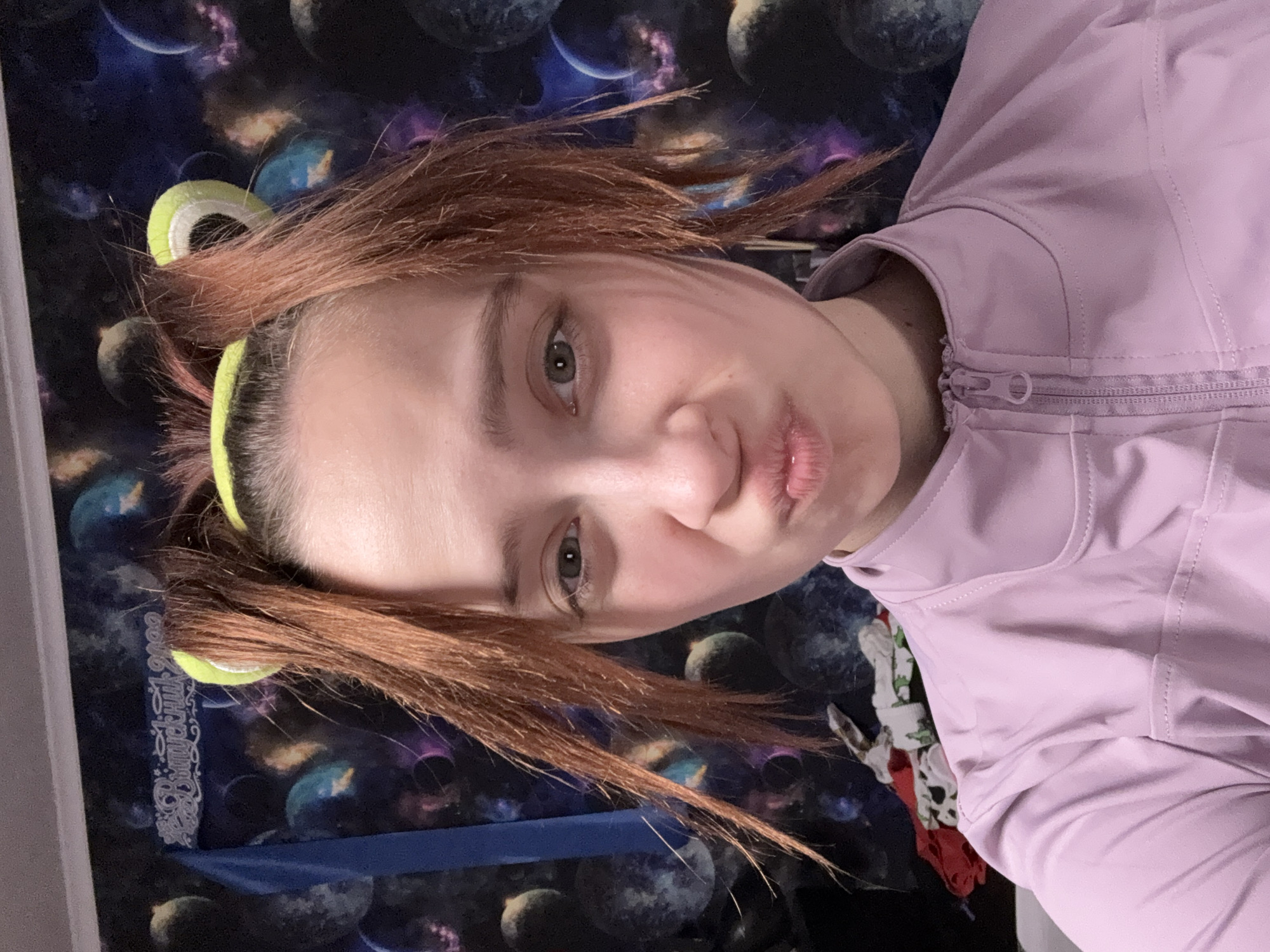 A selfie of a woman with auburn hair is taken indoors against a wallpaper backdrop featuring a galaxy theme of planets and stars. The woman is wearing a pink jacket and a green headband. Her hair is styled in pigtails with distinctive green circular accessories attached near her head. The woman is making a pursed-lips expression. The image does not contain enough information to determine if the location is in Прикамский, Russia.