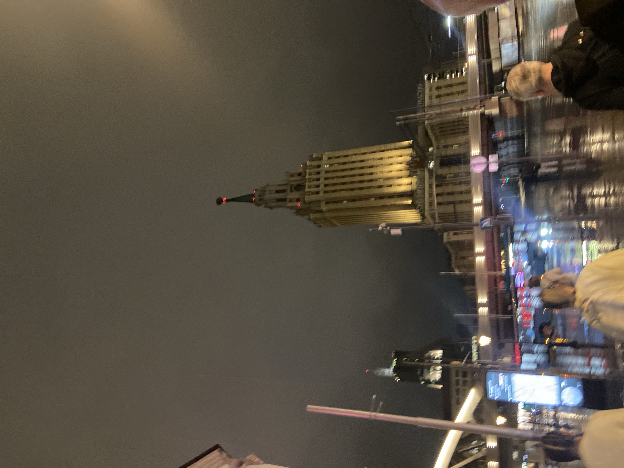At night in Moscow, Russia, a tall building dominates the left side of the frame. The building is lit up with a warm yellow light, while the sky is dark gray. There are other buildings visible in the distance, along with blurred lights. Two people are in the frame: one on the right wearing a black jacket with light hair, and another in the lower right corner wearing a light-colored jacket. They appear to be taking a photo, viewing the screen, or interacting with each other. A metal railing is in the foreground. The scene appears to be taken after rain, with some reflections visible.