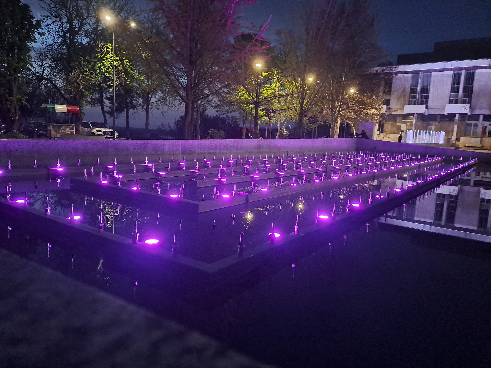 Captured at night, the image presents a water fountain in the city of Unknown, Unknown. The fountain's design incorporates multiple stepped rectangular platforms, each adorned with evenly spaced purple lights. Water is gently spraying upwards from the lights on each platform. The water creates reflections of the purple lights.
In the background, trees with bare branches are illuminated by streetlights, their branches partially obscuring a building with a flat roof and rectangular windows. To the left, a food stall with the colors of the Italian flag is visible, along with a parked car.
Two individuals are discernible in the distance near the building. One is seated, and the other is standing near a traditional water fountain. The sky is dark, suggesting a late evening or nighttime setting.