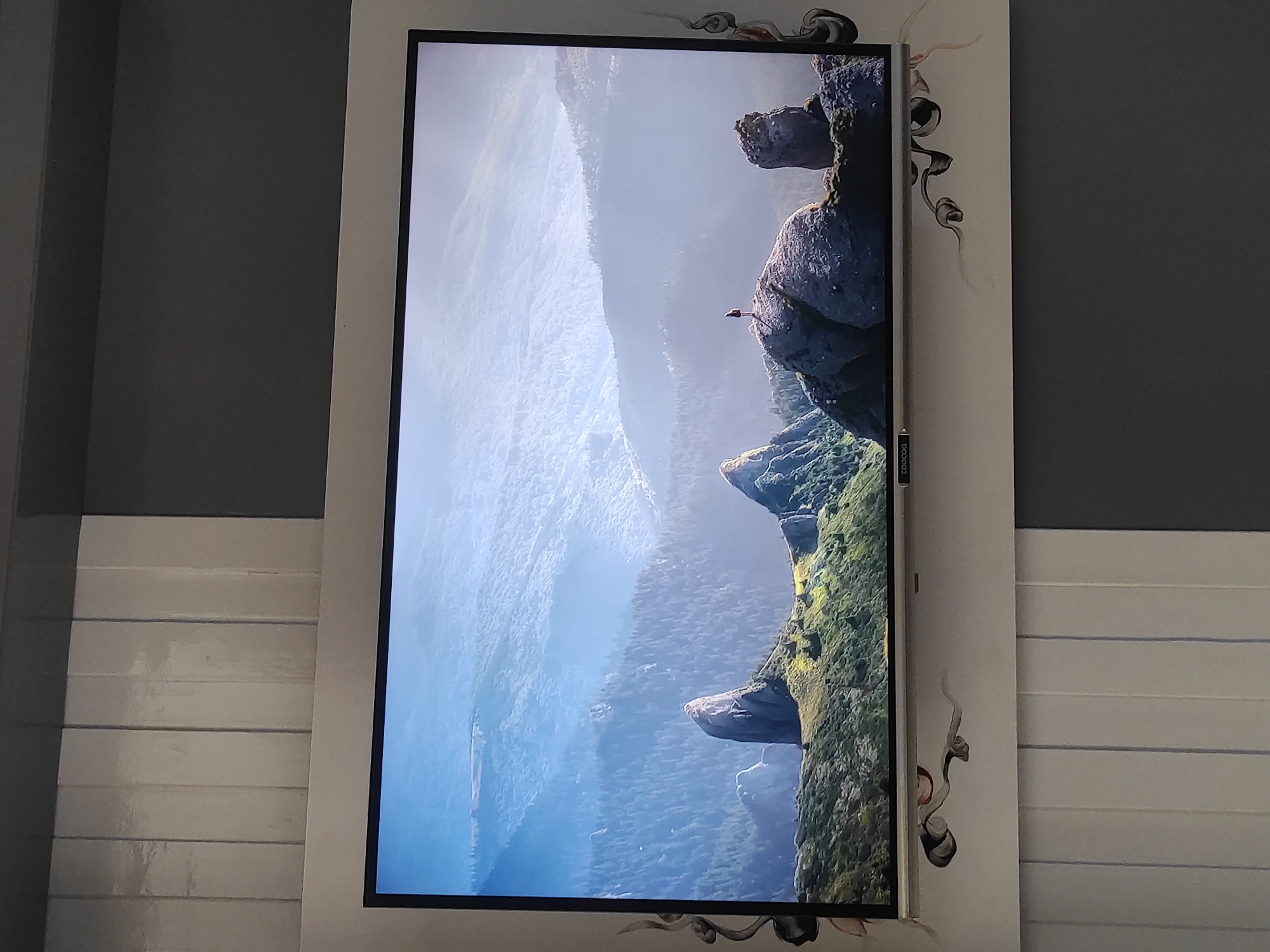 The image shows a television displaying a scenic mountain landscape. The landscape features jagged rock formations covered in greenery, with a misty, layered mountain range in the background. A lone figure stands atop one of the rock formations, silhouetted against the sky. The television is mounted on a wall decorated with vertical white panels and gray sections. Decorative elements resembling swirling lines are attached to the wall below the television. The brand name "COOCAO" is visible on the bottom bezel of the television. The image does not provide any information that would allow for identification of its location in Sumedang, Indonesia.