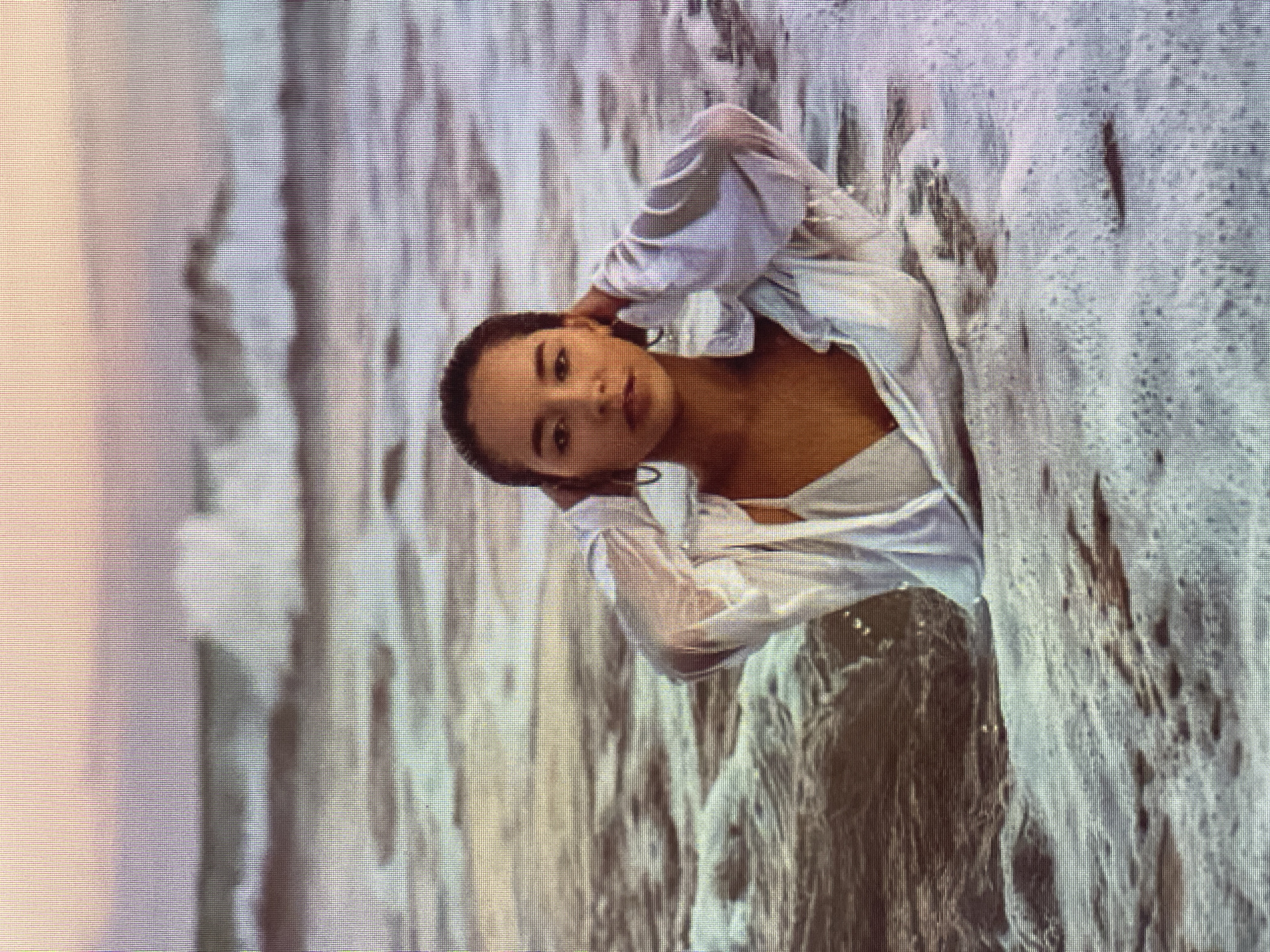 The media file shows a young woman partially submerged in water, with waves and foam surrounding her. She is wearing a white, loose-fitting shirt that appears to be wet. Her hands are raised, with one resting on her head and the other near her neck. She has dark hair and is looking directly at the camera with a neutral expression. The water is a mix of grey and white, suggesting it could be the ocean or a large body of water with some agitation. The lighting suggests it might be daytime, possibly during overcast conditions given the diffused light. The overall impression is a serene and artistic pose, with the water creating a dynamic backdrop.