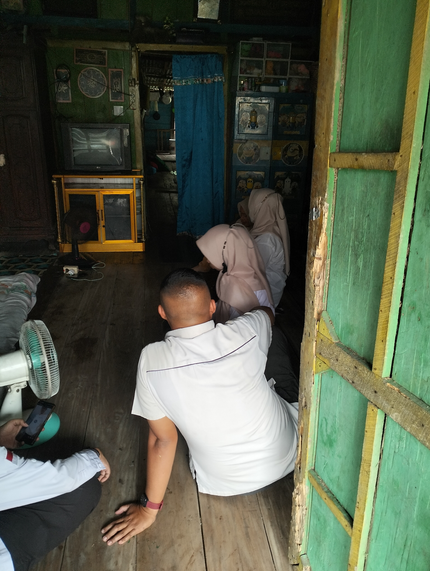 The image depicts an interior scene within a structure located in Mandiangin, Indonesia. The environment appears rustic, with wooden walls and floors. In the foreground, a man wearing a white polo shirt and dark pants is seated on the wooden floor, with his back to the camera. He has a dark watch on his left wrist. To his left, another person, partially visible, is also seated and holding a smartphone. A white electric fan is positioned in front of them.

Further into the room, a television set is visible on a yellow cabinet, with a black fan placed in front of it. Beyond this, two individuals wearing headscarves and white clothing are seated inside an open doorway. The doorway is framed by a green wooden structure. Decorative items, including framed pictures and curtains, adorn the walls. The overall impression is a snapshot of daily life in what appears to be a humble dwelling or community space.