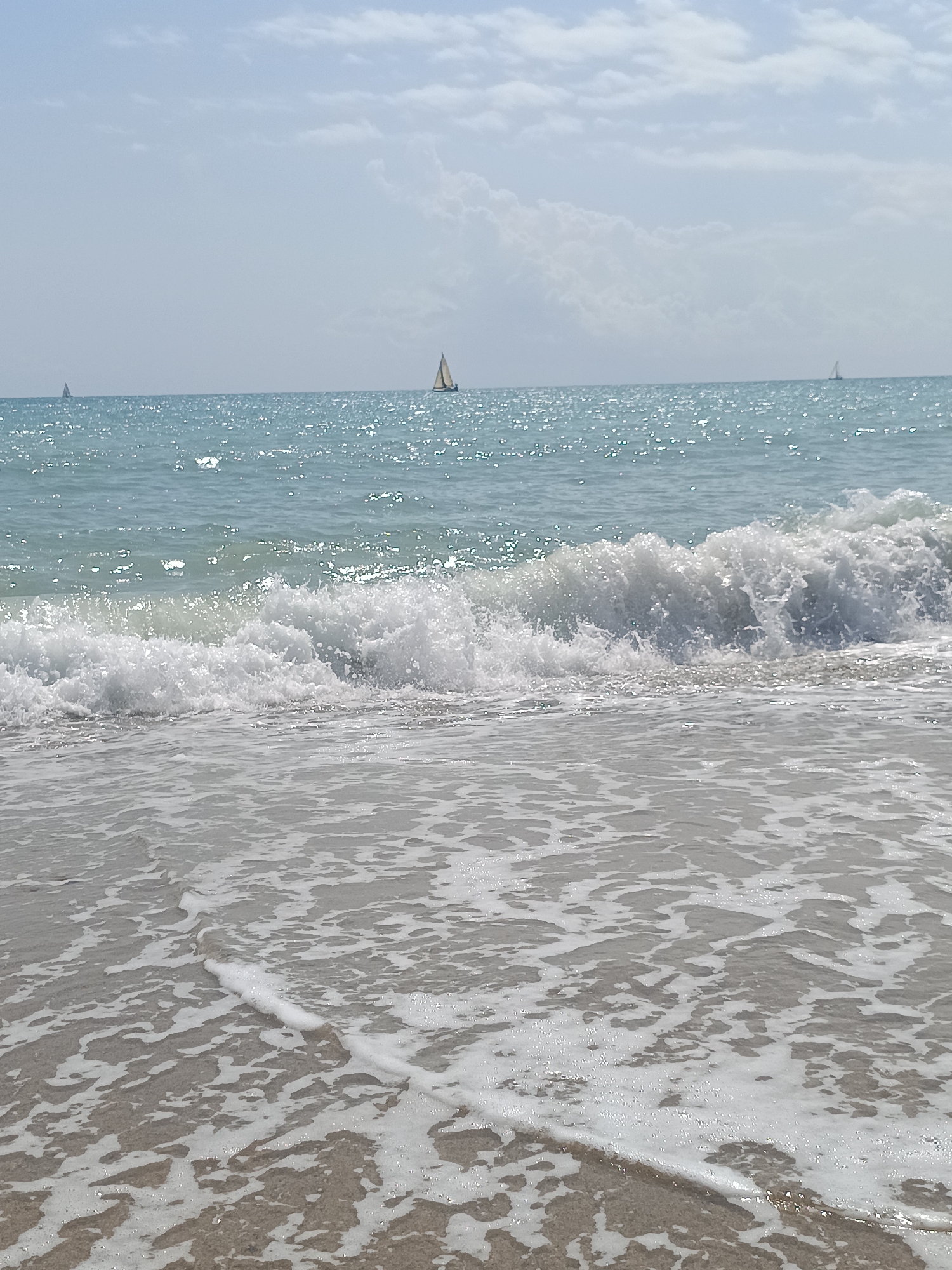A wide-angle, eye-level, vertical shot shows a serene beach scene with two sailboats visible on the horizon. The water is a vibrant turquoise, reflecting the bright sunlight, and small waves are breaking closer to the shore, creating white foam that washes onto the sandy beach. The sky is a pale blue with scattered white clouds. The foreground is dominated by the foamy water and sand. The scene is tranquil and evokes a sense of peace and relaxation. The location is the beach of Sagunto, Spain, a coastal city known for its historical significance and beautiful Mediterranean coastline.