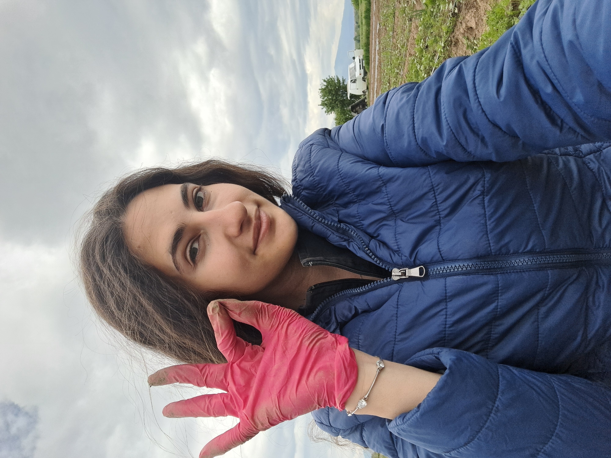 A young woman is pictured in a selfie taken in a field in Negovan, Bulgaria. She is wearing a blue puffer jacket and a pink glove on her left hand, which is raised to display some dirt. A silver bracelet adorns her wrist. The background reveals a cloudy sky above a field with a white van and green trees visible in the distance.