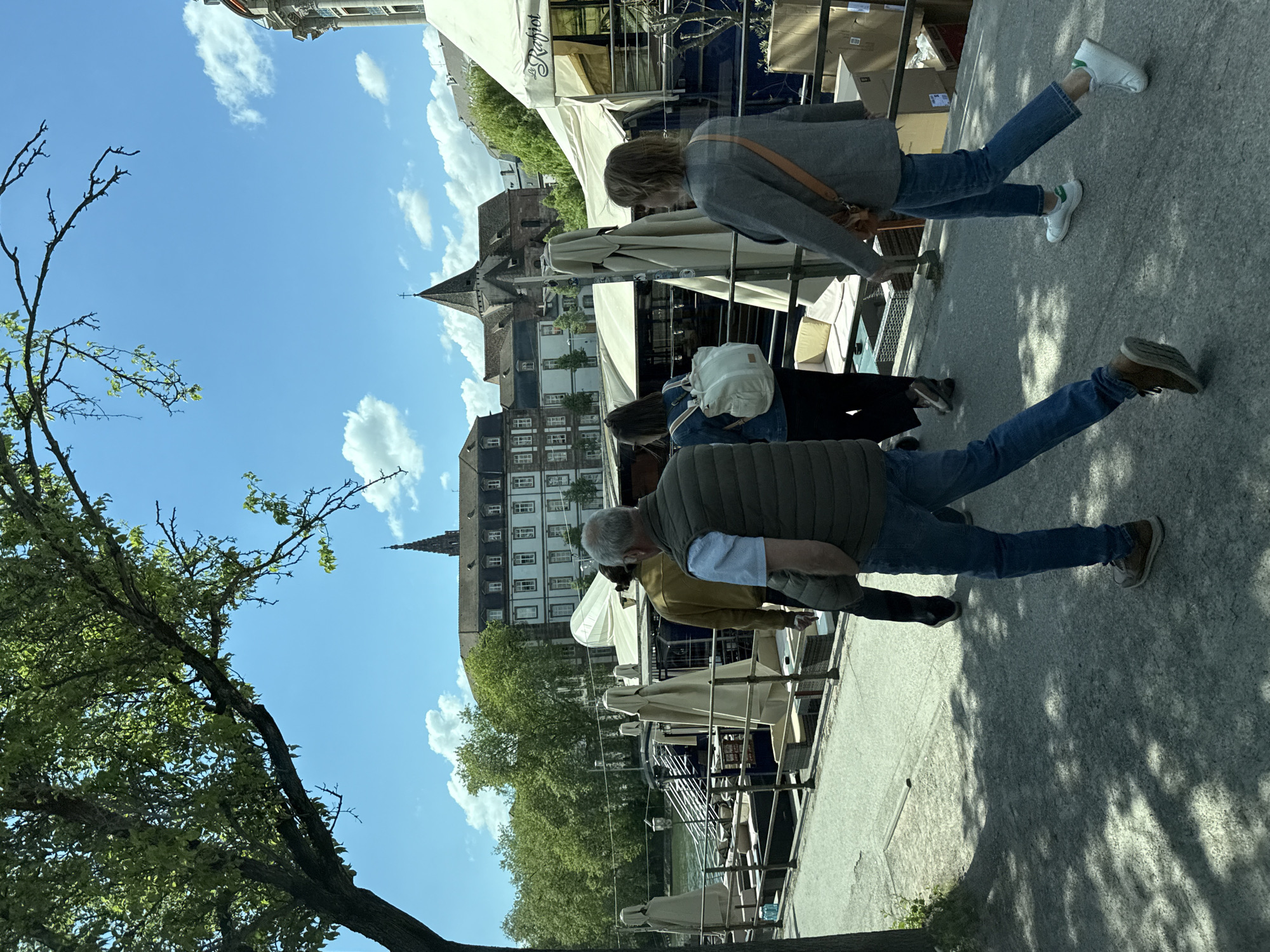 The image depicts a vibrant daytime scene along a waterfront promenade in what appears to be a European city, likely Strasbourg, France, given the architectural style. The perspective is slightly low, looking towards a row of historic buildings and a prominent church spire under a bright blue sky with scattered white clouds.
In the foreground, two individuals are walking away from the viewer on a paved path. One person on the right wears a grey jacket, blue jeans, and white sneakers with green heel tabs. The other, slightly behind and to the left, wears a striped vest, light-colored shirt, blue jeans, and brown shoes. Dappled shadows, likely from the large tree on the far left with green foliage, fall across the path.
Along the waterfront, several boats or floating platforms are moored, serving as outdoor dining areas. These are covered by large, light-colored awnings or umbrellas. One awning clearly displays text that appears to be "Le Rappot". Further back, other people can be seen near these establishments, including one person with a light-colored backpack.
The background features classic European architecture, including multi-story buildings, some with visible windows and intricate facades. A tall, dark church spire with a cross on top rises above these buildings. The general atmosphere suggests a pleasant, sunny day conducive to strolling and outdoor leisure activities.