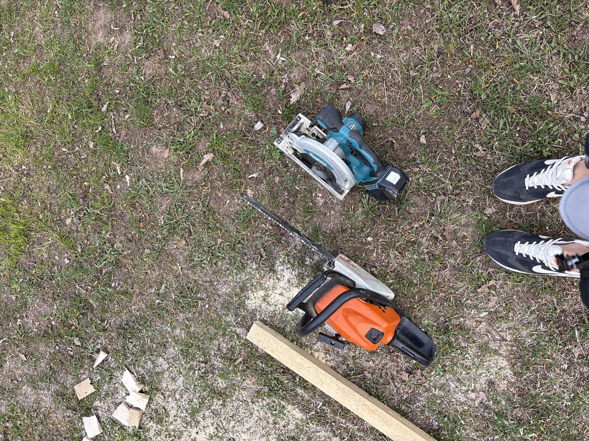 This overhead shot, captured in Chaikovsky, Russia, showcases a ground-level scene featuring woodworking tools and materials. The ground is covered with sparse green grass mixed with patches of brown dirt and scattered dry leaves.
In the upper-middle section of the image, a teal and black Makita cordless circular saw, with its battery attached, rests on the ground. Below it, an orange and black chainsaw with a visible metal bar and chain also lies on the dirt. A light-colored wooden plank or beam is partially visible at the bottom, extending horizontally. Numerous small, irregular pieces of cut wood are scattered near the bottom left, surrounded by a substantial amount of fine sawdust, indicating recent cutting activity.
On the far right side of the frame, the lower parts of a person's legs and feet are visible. The person is wearing black sneakers with white laces and soles, along with a light-colored sock or ankle cuff.
The scene suggests an outdoor work environment, likely involving cutting wood for construction, renovation, or yard maintenance. The presence of both a circular saw and a chainsaw implies a variety of cutting tasks. The time of day appears to be daylight, and the overall impression is one of active work or a temporary pause in it. The text "Makita" is clearly visible on the circular saw.