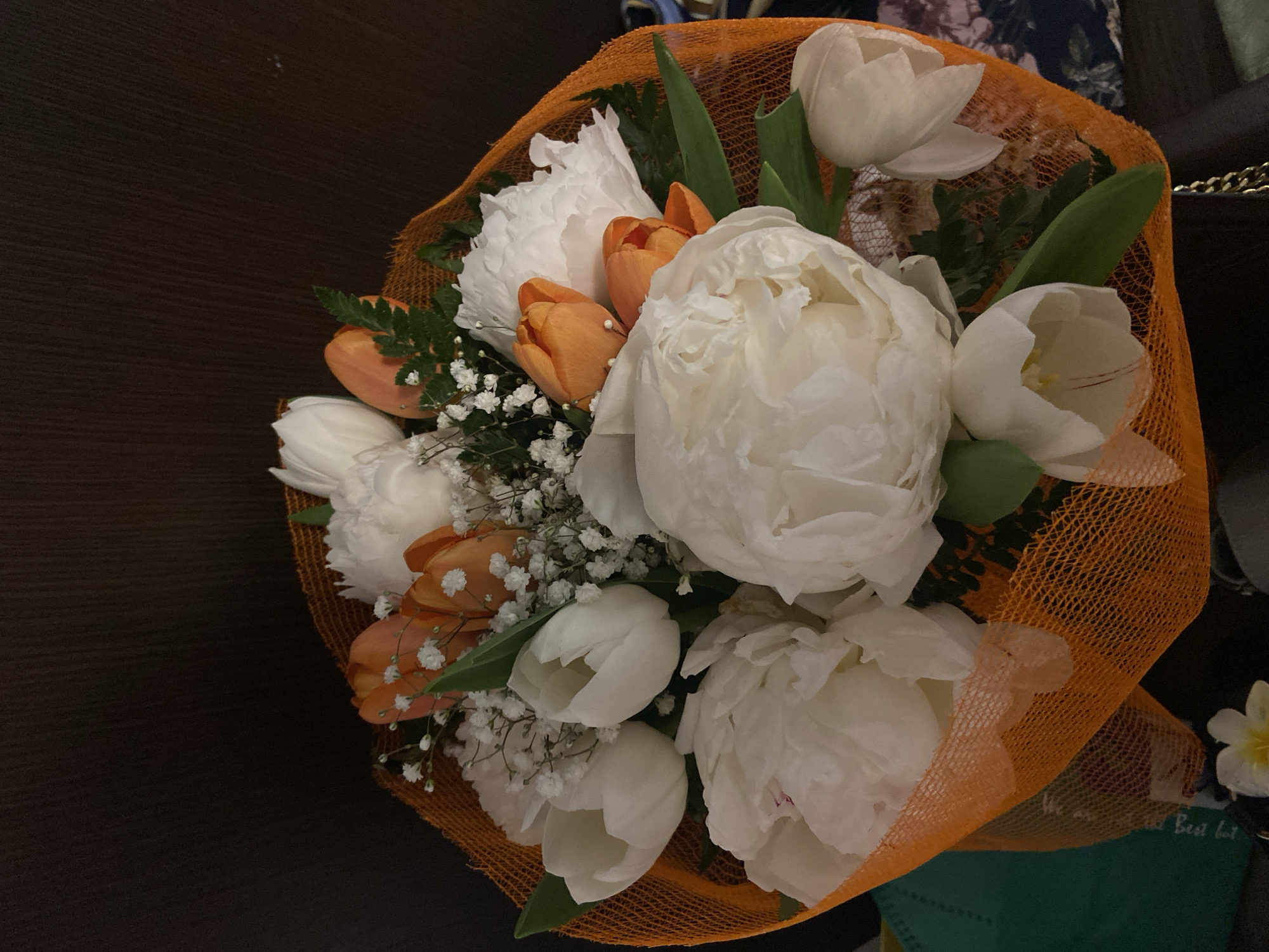 A bouquet of flowers rests on a dark, possibly wooden, surface. The bouquet is wrapped in orange netting. It includes large white peonies, white tulips, and orange tulips. There are also smaller white filler flowers, possibly baby's breath. The flowers are interspersed with green leaves. The overall composition is rich in color and texture. It is not possible to determine the location in the city of Lecco, Italy.