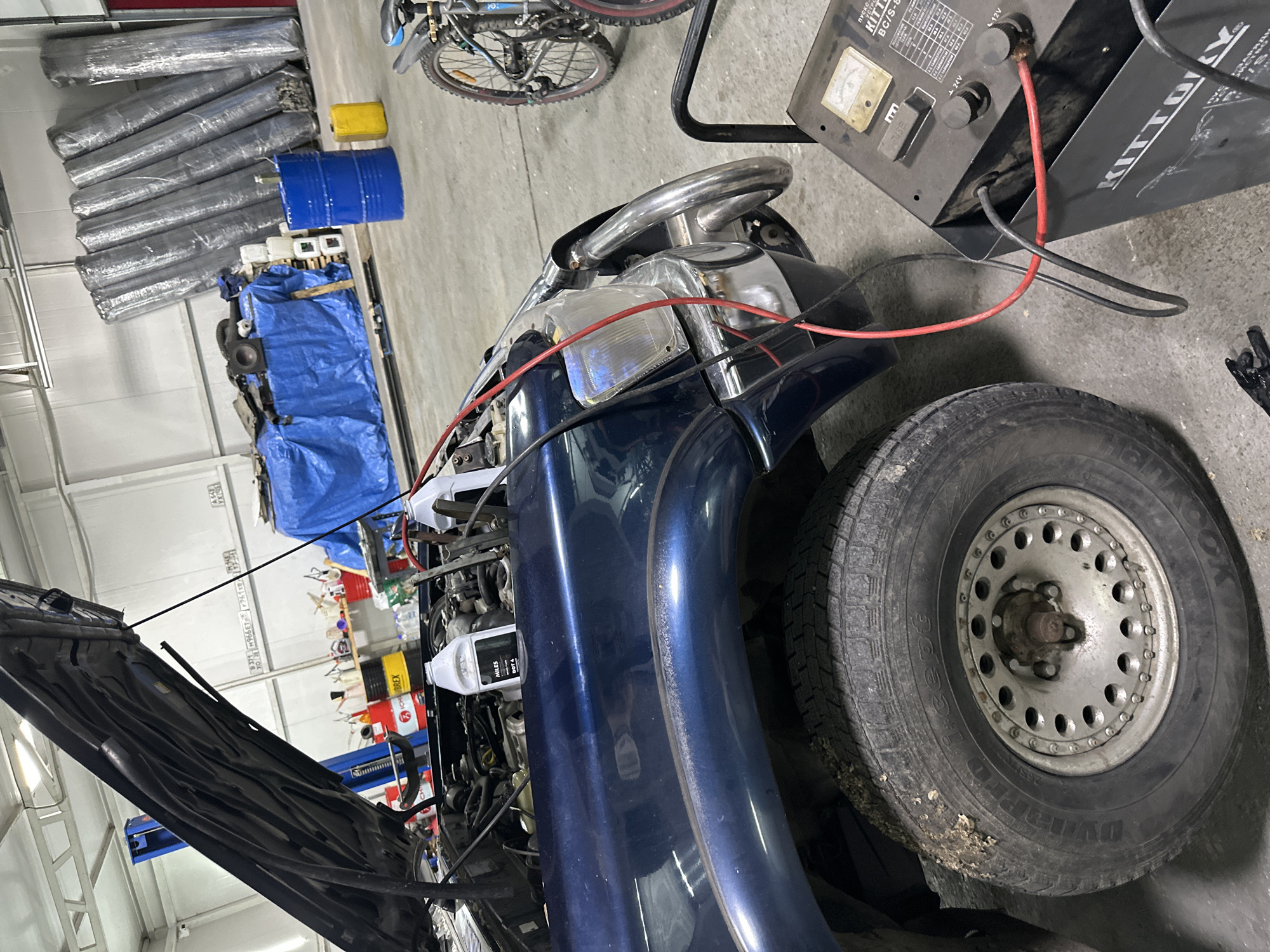A dark blue vehicle with its hood open is parked in a garage. The car's engine is visible, along with a white bottle of fluid labeled "MILES DOT 4". A battery charger is connected to the car with red and black cables. In the background, there are stacks of rolled-up material, several barrels, and a bicycle. The scene appears to be a vehicle repair or maintenance setting, possibly in a commercial garage. No people are visible in the image. The image does not provide any specific information about the city of Blagoveshchensk, Russia.