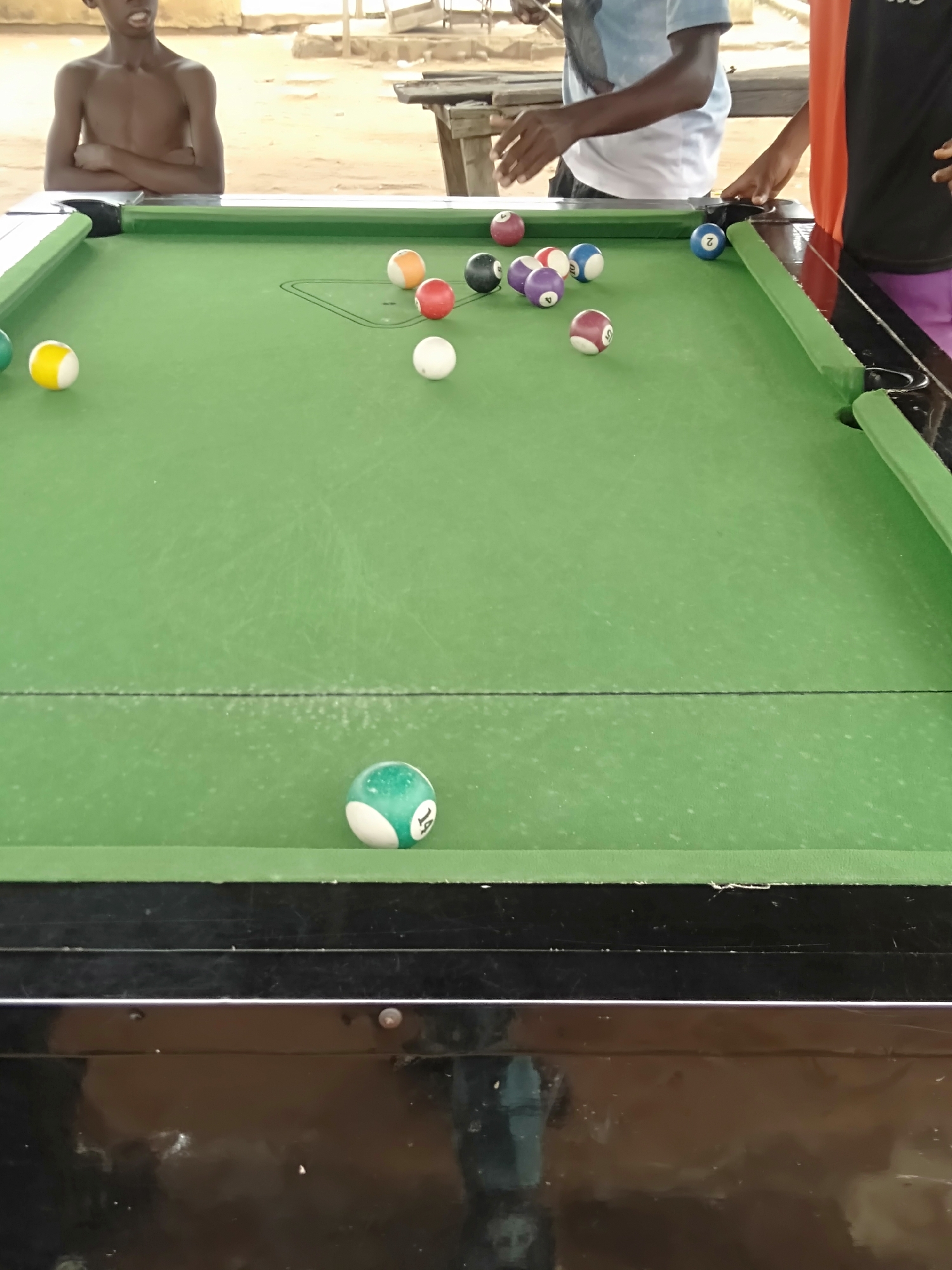 The image shows a billiards table with multiple pool balls scattered across its green surface. At the far end of the table, pool balls are racked, including balls of various colors such as orange, red, black, purple, and blue. To the left of these, there is a single yellow ball near the edge of the table. Further down the table, toward the front, a green ball marked with the number "14" is positioned near the edge.
Three people are partially visible around the table. A person in a light blue shirt is seen near the racked balls with their hands near the table surface. Another person is standing near the right edge of the table, wearing an orange top over a black shirt and purple pants. A third person, shirtless, is positioned near the left side of the table, facing forward with arms crossed.
The table itself is black with green felt and is situated in an outdoor environment. There is a sandy area visible in the background. Wooden structures are also visible, possibly indicating a makeshift or informal setting.
The image does not provide enough information to identify the city or location in the city of Unknown, Unknown.