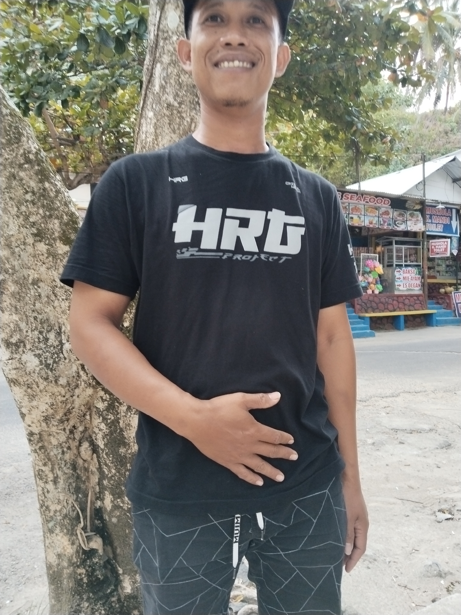 A medium shot captures a smiling Indonesian man standing outdoors, leaning against a textured tree trunk. He is wearing a black t-shirt with a white graphic logo that reads "HRG Project" and black shorts with a geometric pattern. His left hand is resting on his stomach. In the background, a street scene unfolds with a building displaying signage for "SEAFOOD", "MUSOLA K. MANDI TOILET", and food items like "BAKSO MIE AYAM ES DEGAN". Palm trees and foliage are visible, suggesting a tropical or coastal environment. The ground appears to be paved or a dirt road. The overall impression is a casual, everyday moment likely captured in Prigi, Indonesia, given the context provided by the background signage.