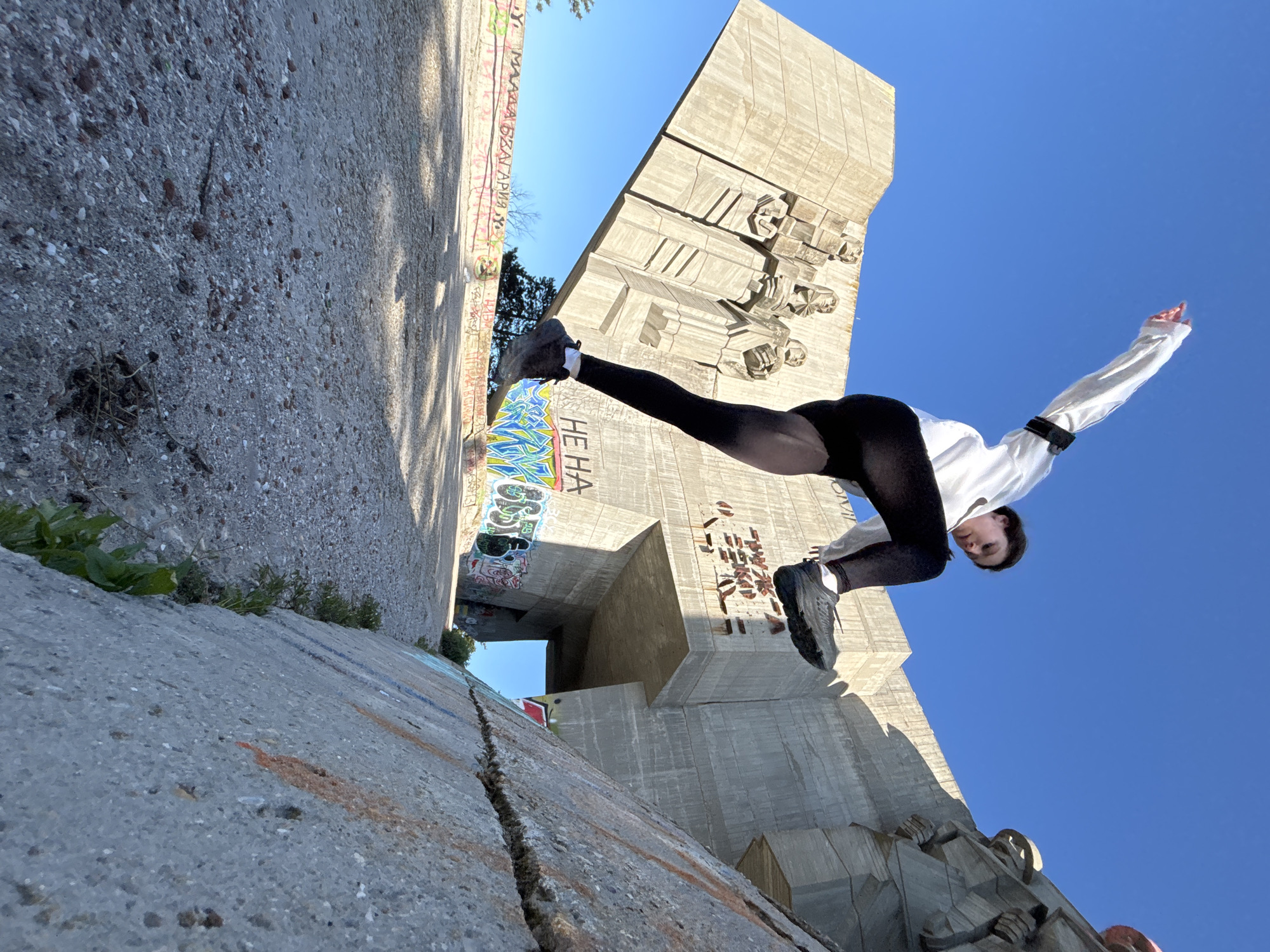 A dynamic low-angle shot captures a person in an athletic pose against the backdrop of a massive concrete monument in Varna, Bulgaria. The image is rotated approximately 90 degrees counter-clockwise, making the ground appear as a vertical wall on the left, and the monument seemingly leaning.
The primary subject is a young woman, seen from below, mid-action. She wears black leggings, dark sneakers, and a long-sleeved white top, possibly a cropped shirt or jacket. Her left leg is extended high, bent at the knee, while her right leg is firmly planted. Her arms are outstretched, one slightly raised, contributing to a sense of balance and movement, resembling a high kick or a dramatic dance jump. Her head is tilted slightly down, and her expression is focused.
The background is dominated by a large, brutalist concrete monument featuring imposing bas-relief sculptures of human figures, likely depicting historical or heroic themes. The monument's surface is textured and shows signs of age. Above the monument, a clear, vibrant blue sky suggests a bright, sunny day. To the left of the person, a concrete wall, which in reality would be the ground or a platform, is visible. This surface is rough, composed of speckled aggregate, and displays cracks and patches of green vegetation. Graffiti in various colors, including blues, yellows, reds, and black, adorns the concrete surfaces, adding a contemporary, urban layer to the historic setting. Visible text fragments include "HE HA" on a graffiti piece near the person's extended leg, and other Cyrillic script on a distant wall, such as partial words resembling "МАЛКА БЪЛГАРИЯ" (Small Bulgaria).
The strong natural light casts distinct shadows, highlighting the architectural details of the monument and the textured surfaces. The unusual photographic angle emphasizes the scale of the monument and the dynamism of the person's pose.