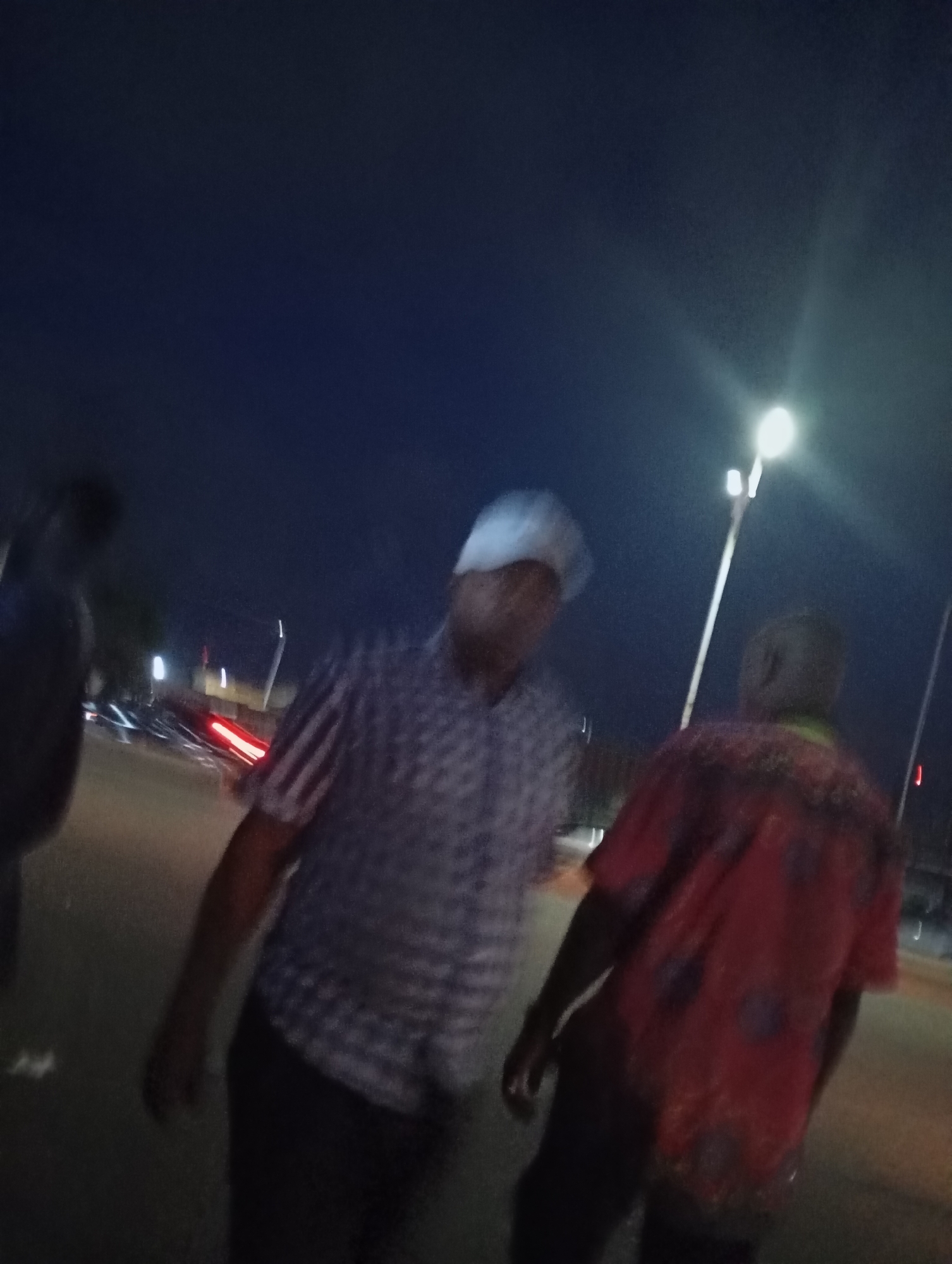 The image shows a nighttime scene on a street in Jos, Nigeria. The sky is dark blue, and a bright streetlight illuminates the area. Several people are visible, though their features are blurred due to the low light conditions and motion.
In the foreground, a person wearing a light-colored cap and a patterned shirt is walking. To their right, another person is visible from the back, wearing a red shirt with a pattern. Other blurred figures and vehicles, including ones with visible red taillights, suggest a street or road environment. The background shows some indistinct structures and the dark sky. The overall impression is of a casual nighttime gathering or movement on a street.