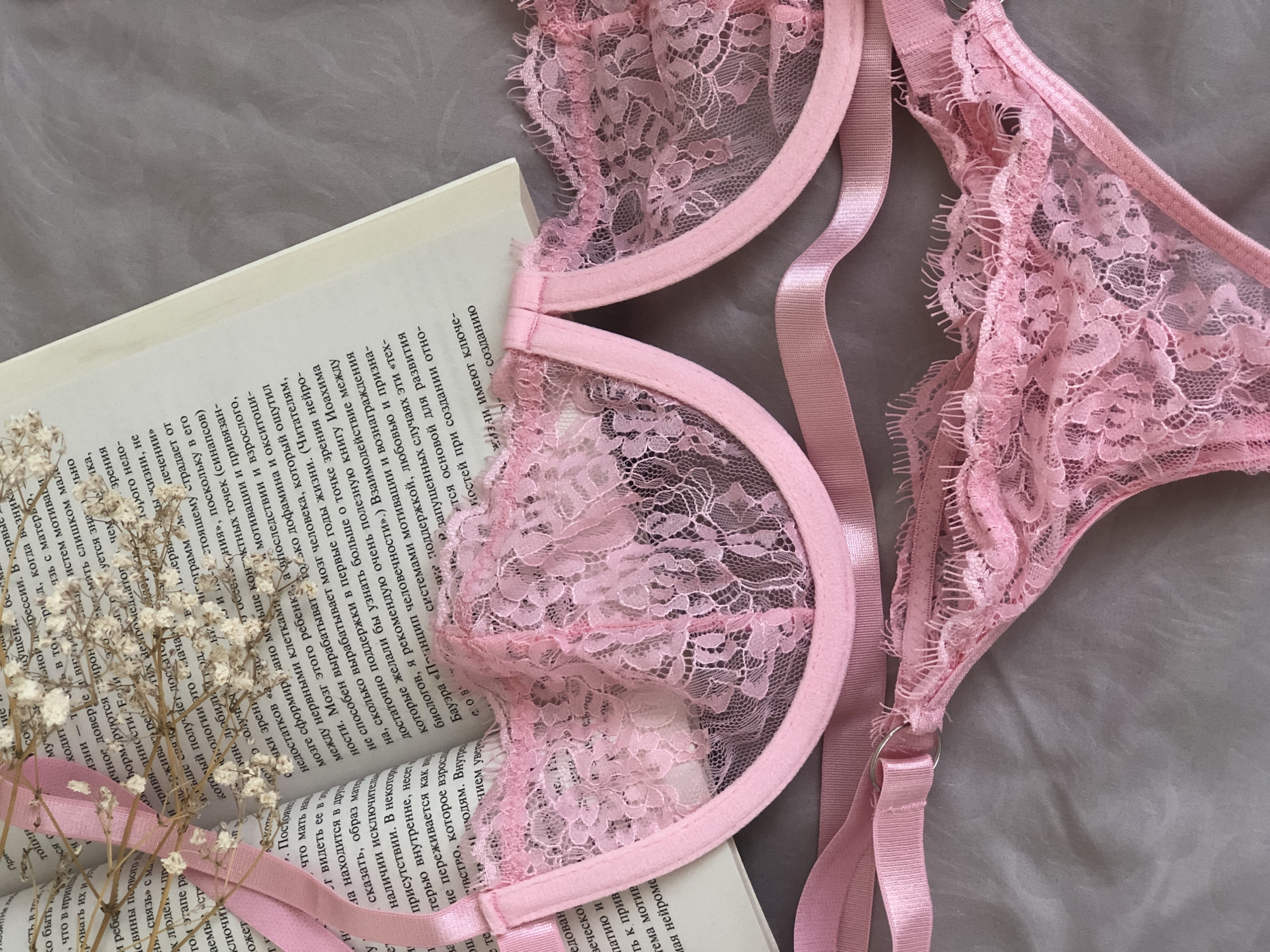The image is a flat lay, focusing on a pink, lacy bra and panties set laid atop an open book and a soft grey surface. The bra is prominently displayed, with its delicate lace detailing and pink straps partially covering the book's pages. The open book lies beneath a portion of the bra. Some baby's breath flowers lie on the book. The grey surface serves as a neutral backdrop. The scene suggests a feminine, intimate, and possibly romantic or sensual setting. There is no city in the image.