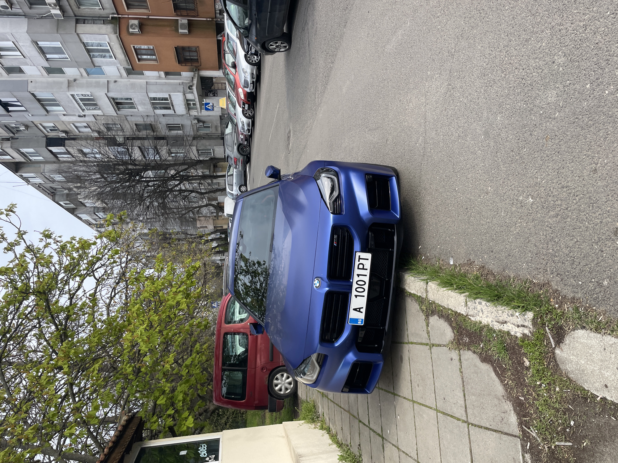A blue BMW M2 with Bulgarian license plate "A 1001 PT" is parked on the street next to a sidewalk in an urban area. The car is adjacent to a red van. Behind the BMW, there is a row of parked vehicles. In the background, there is a block of buildings with numerous windows and air conditioning units. A leafless tree is visible near the building. The foreground includes a sidewalk with some vegetation growing along the edge. Based on the license plate information, the location is likely in the city of Burgas, Bulgaria. There are no people visible in the image.