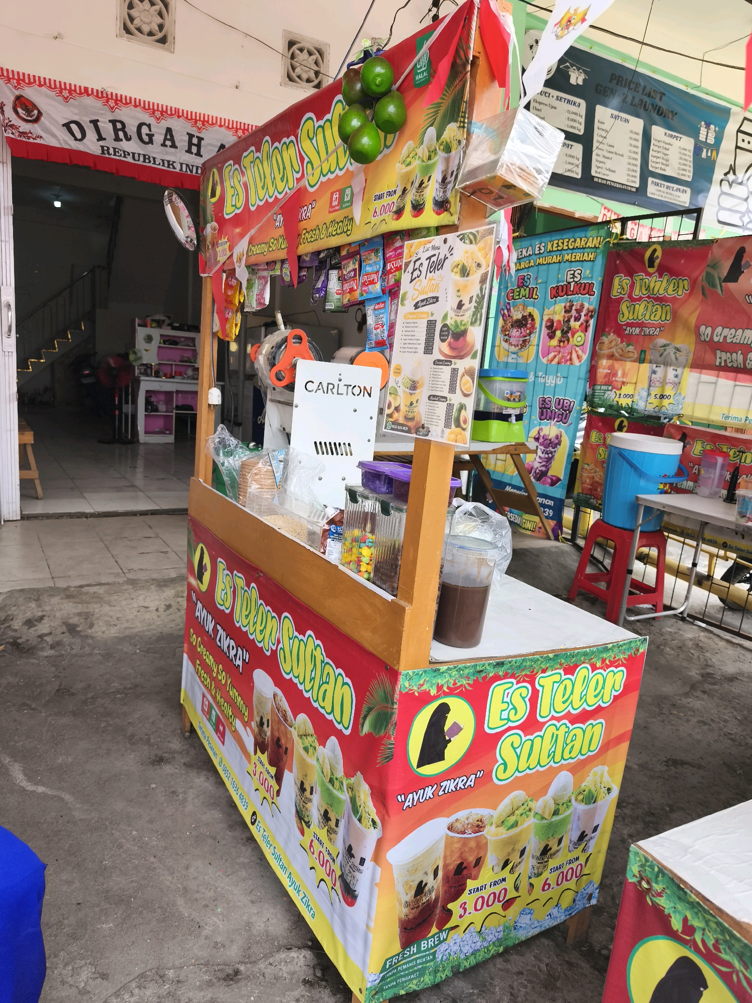 The image depicts a street-side food cart selling "Es Teler Sultan" (Sultan's Avocado Ice) and other beverages. The cart is brightly decorated with red and yellow banners featuring images of the drinks and prices. In the background, an open doorway leads into a building, revealing a staircase and a display of various merchandise. A banner above the doorway reads "DIRGAHAYU REPUBLIK INDONESIA" (Happy Indonesian Republic Day), indicating the patriotic context. The scene appears to be in Palembang, Indonesia, given the presence of Indonesian text on the banners and the architectural style visible. The cart is equipped with a drink dispenser, cups, and various ingredients. There are no people actively engaged in transactions, but the cart is set up for business. The surrounding environment suggests an urban setting with concrete ground and adjacent buildings.