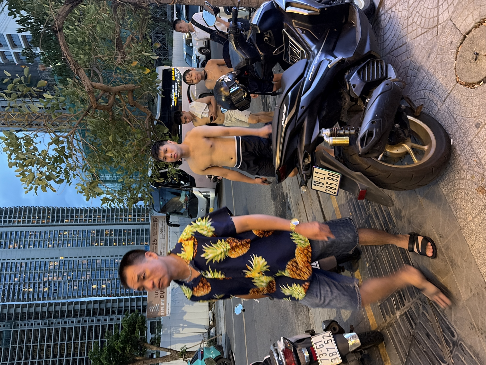 In Đà Nẵng, Vietnam, a group of young men are walking on a sidewalk next to a street at dusk. The man in the foreground, wearing a dark blue short-sleeved shirt with a pineapple print and denim shorts, walks with his left leg extended forward. To his right, another shirtless young man wearing black shorts stands with his hands at his sides. Behind him, another shirtless young man is holding a towel. Further back, another young man in a white shirt with a black bag slung over his shoulder is visible. Several motorcycles are parked along the sidewalk, one of which is in the foreground, a black scooter. In the background, tall modern buildings loom, and a bus is parked on the street. The sky is a dim blue, indicating twilight.