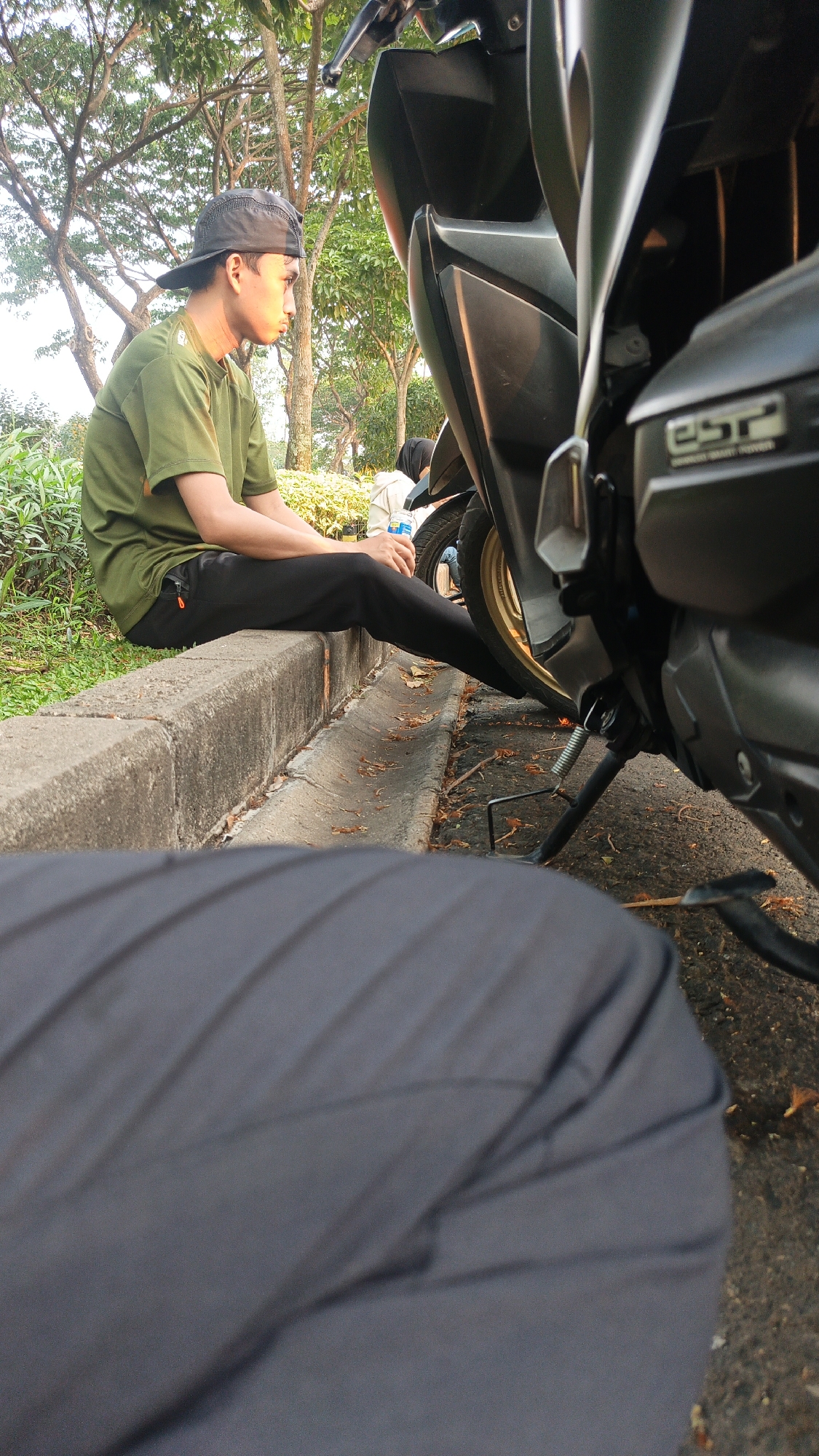 An outdoor scene shows a young male wearing a dark baseball cap and an olive green t-shirt, sitting on a concrete curb. He is facing to the right, looking away from the camera. Black pants with an orange zipper are visible on his legs. Behind him, a dark-colored scooter is parked. The scooter's front portion is in the foreground, angled towards the right. The background features lush green trees and foliage, suggesting a park or roadside setting. The ground is paved with asphalt and scattered with fallen leaves. A small ditch runs alongside the curb. The image appears to be taken in Bacin, Indonesia, as indicated by the context.