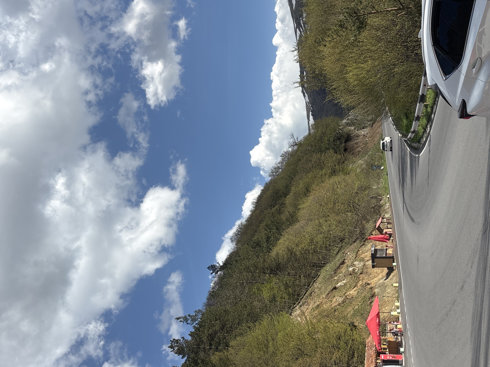 The image captures a winding road in Dilijan, Armenia, bordered by a steep, tree-covered hillside and a cloudy sky. A white car is visible on the right edge of the frame, partially cut off. A smaller, white vehicle is further down the road. Along the hillside, there are small rest stops with red umbrellas. The sky is a mix of blue and fluffy white clouds.