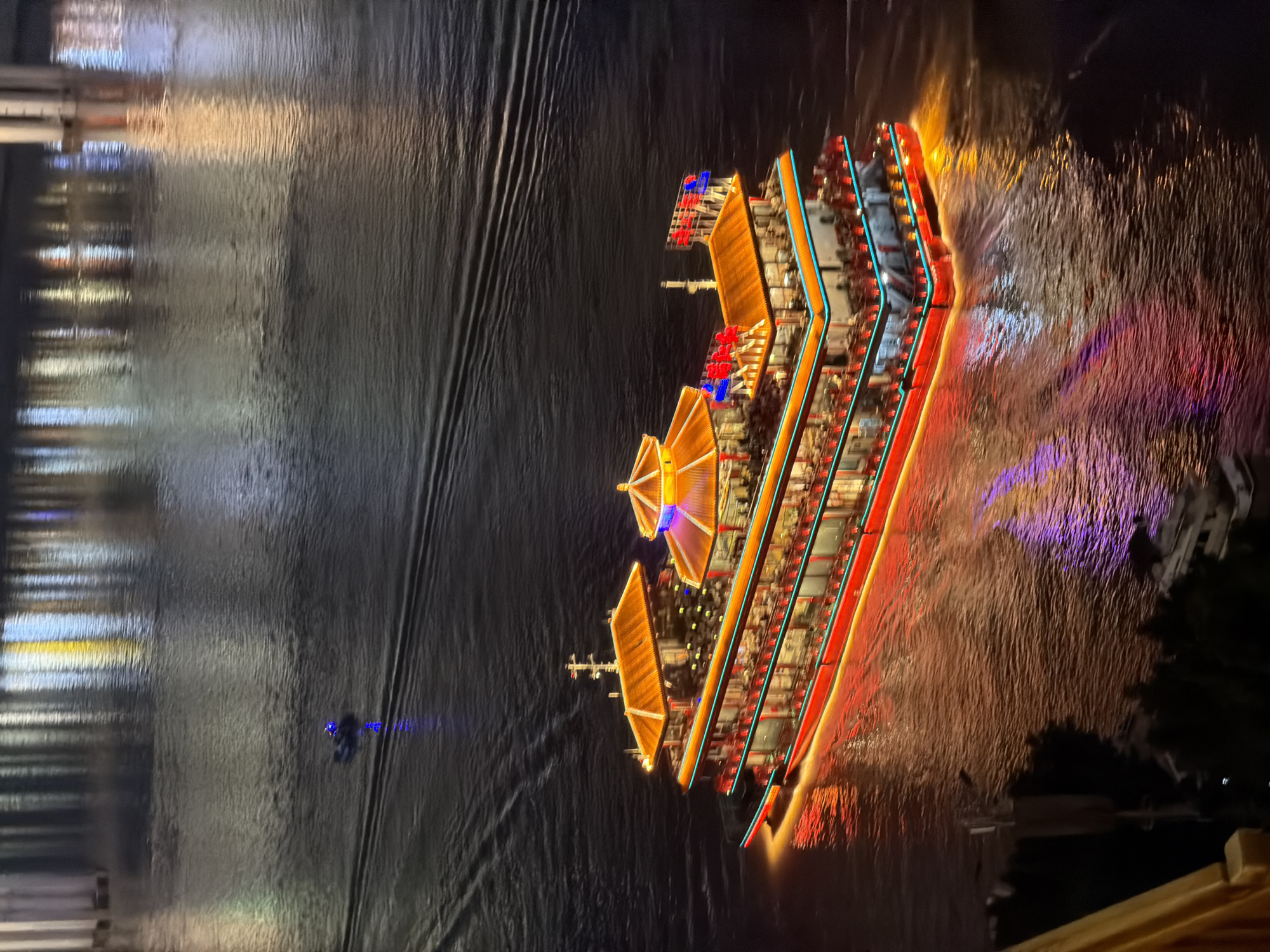 This is a high-angle, eye-level, medium shot of a brightly lit, multi-decked boat navigating a dark body of water at night. The boat, likely a tourist vessel, features traditional Chinese architectural elements with ornate, orange-tiled roofs and vibrant red and blue LED lighting illuminating its exterior. Numerous small red lights are visible along the railings and windows of each deck, suggesting the presence of many people inside. The boat is leaving a wake in the water, indicating movement.
In the distance, a small, dark vessel with blue lights is also visible, leaving a similar, though fainter, wake. The water reflects the lights from the boat and the distant cityscape, creating streaks of orange, yellow, purple, and blue on its surface. The background shows blurred reflections of lights from buildings or bridges, characteristic of an urban waterfront environment. The overall scene suggests a lively atmosphere on the water, likely within a city known for its nighttime illuminations and river cruises. Given the style of architecture and the typical tourist attractions in China, this scene is likely located in the Yuzhong District of China, known for its riverside developments and night tours.