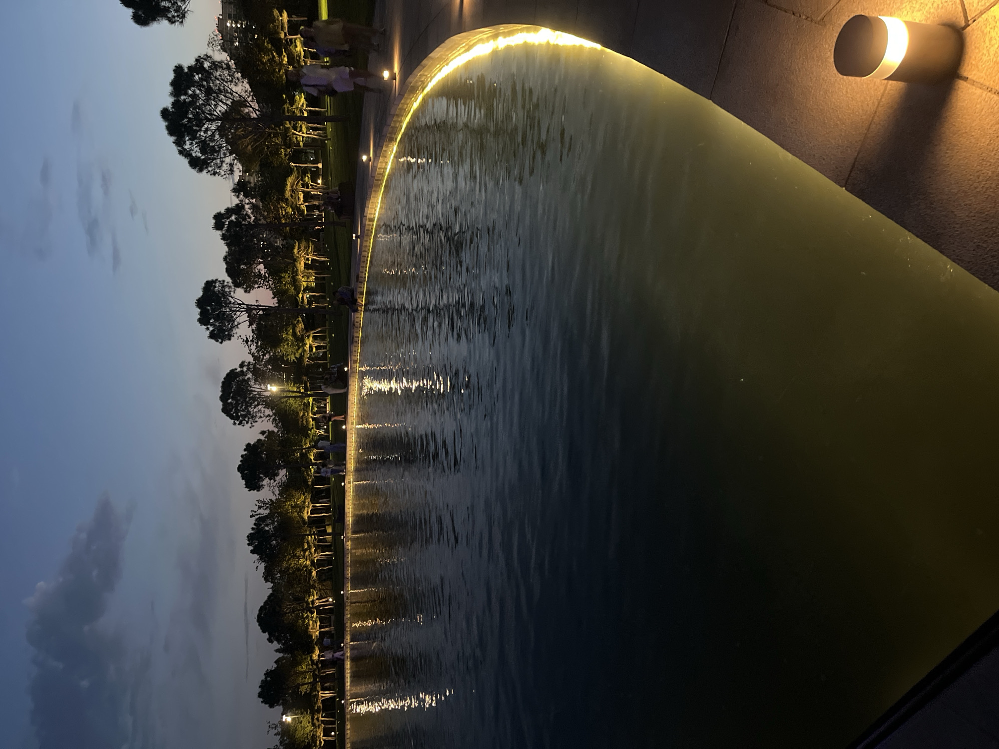 The image shows a park scene at dusk, likely in Krasnodar, Russia. The perspective is low and tilted, focusing on a water feature that appears to be a decorative pool or fountain. The water's surface is rippling, reflecting the soft, warm glow of lights lining the edge of the pool and the pathway beside it. These lights create a shimmering effect on the water and illuminate the surrounding area.
Beyond the water feature, a line of trees with lush green foliage stands tall, their silhouettes becoming more defined against the darkening sky. Behind the trees, several small figures of people can be seen walking or standing along the pathway. They appear to be casually enjoying the evening ambiance of the park. A few small, cylindrical lights are mounted on a stone structure to the right of the frame, casting warm light onto the textured surface. The sky is a deep blue with hints of fading daylight, and a few wispy clouds are visible. The overall mood is serene and peaceful, suggesting a pleasant evening stroll in an urban park.