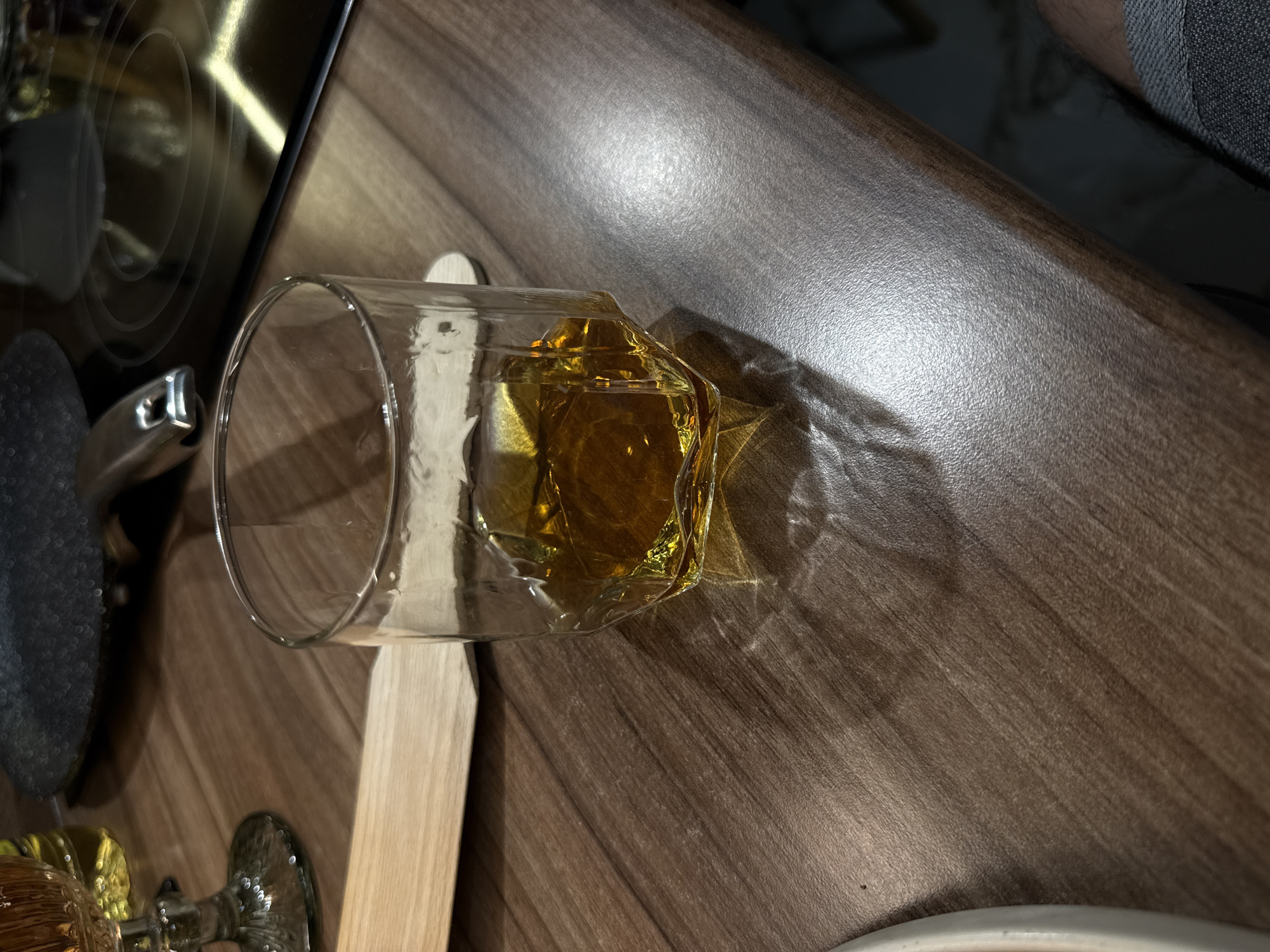 A clear glass containing an amber liquid, likely whiskey or a similar spirit, lies on its side on a dark wooden surface with a shiny, light-reflecting finish. A light-colored wooden stirring stick rests beneath the glass. To the left, a portion of a black stovetop is visible, with a metal handle and a dark-colored pan or cooking surface. In the lower-left corner, a portion of a glass bottle with a decorative, ornate stopper can be seen. The liquid has spilled out of the glass onto the surface, creating a transparent stain. The background shows a glimpse of a white tile floor and part of a person's leg with dark-colored clothing.
The scene is indoors, possibly in a kitchen or bar setting. Based on the style of the furniture and the overall ambiance, it's impossible to pinpoint a specific location in Sofia, Bulgaria, without more information or context.