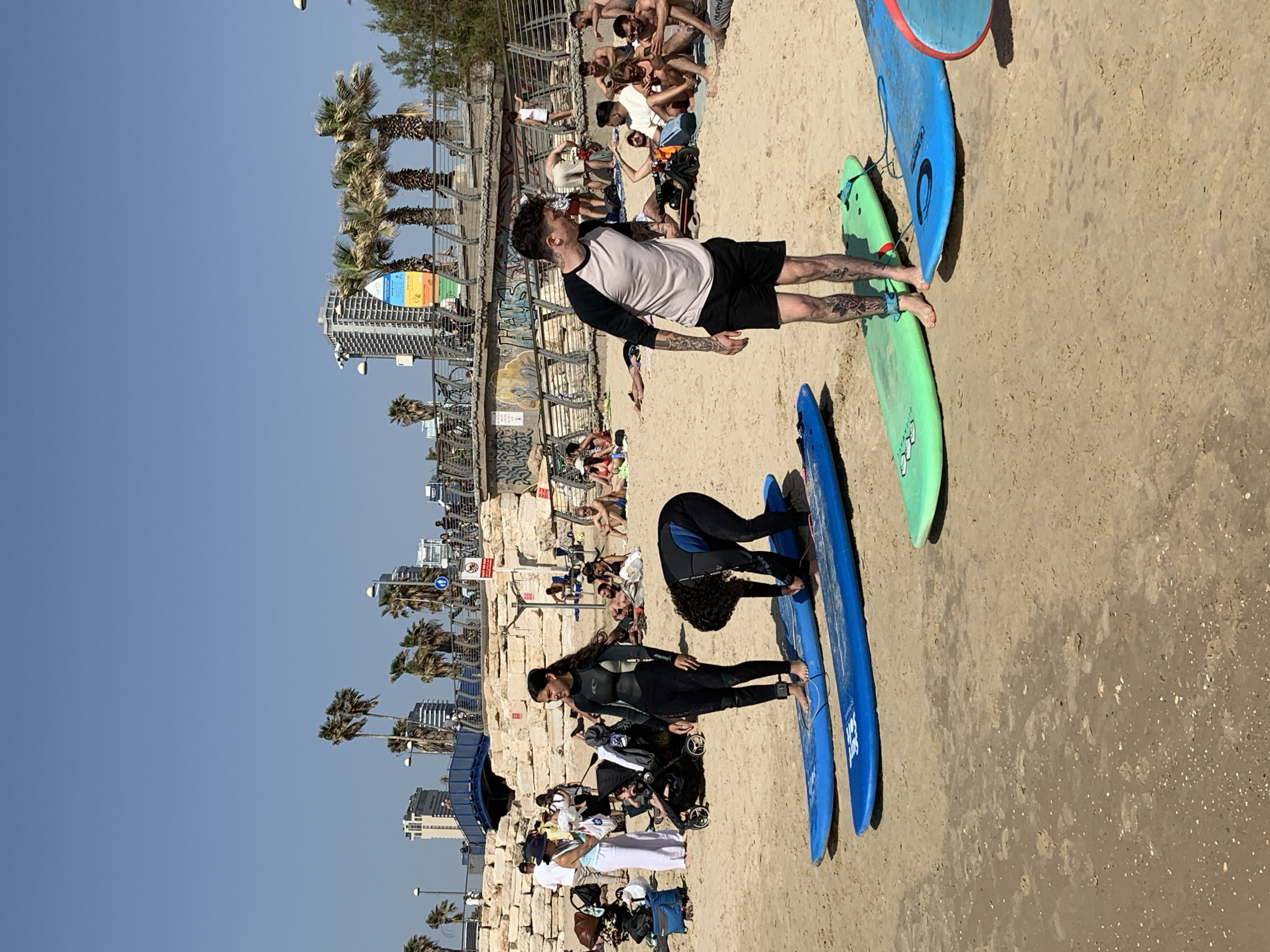 The image shows a sunny beach scene in Tel Aviv, Israel, likely along the Mediterranean coastline, as suggested by the presence of modern buildings and palm trees visible in the background. The foreground of the beach is sandy with several surfboards lying on the sand.
Several people are present in the scene. Two individuals wearing wetsuits stand next to blue surfboards, appearing to be preparing for surfing or receiving instruction. Nearby, another person in casual clothing (a light-colored shirt with black sleeves and dark shorts) is standing on the sand, with visible tattoos on their legs and arms.
A group of people are gathered near a low stone wall and a metal railing, situated between the beach and the promenade. Some appear to be sunbathing or relaxing. Additionally, there is a baby stroller with several bags nearby.
The general atmosphere is relaxed and beach-oriented, capturing a typical scene of leisure activities in an urban coastal environment.