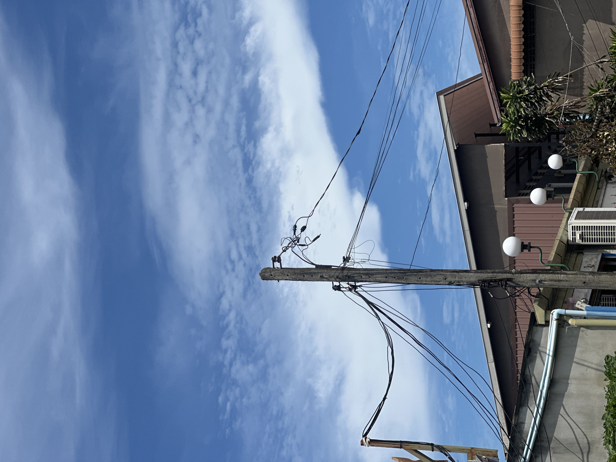 In Unknown, Unknown, a concrete utility pole with multiple wires is positioned in the foreground against a bright blue sky with scattered white clouds. The pole is weathered and carries a complex network of black electrical wires. To the right of the pole, a white, globe-shaped light fixture is attached to a green bracket. Behind the utility pole and wires, a multi-level building complex is visible, with the top sections featuring brown roofs and angled eaves. Some sections of the building are clad in corrugated metal. A white air conditioning unit is mounted on a lower level of the building, to the right of a gray wall with visible pipes. Green foliage is present at the bottom left of the frame, and more dense greenery with thin trunks is visible to the right, partially obscuring the lower parts of the buildings. The scene appears to be outdoors during the daytime, with clear weather conditions.