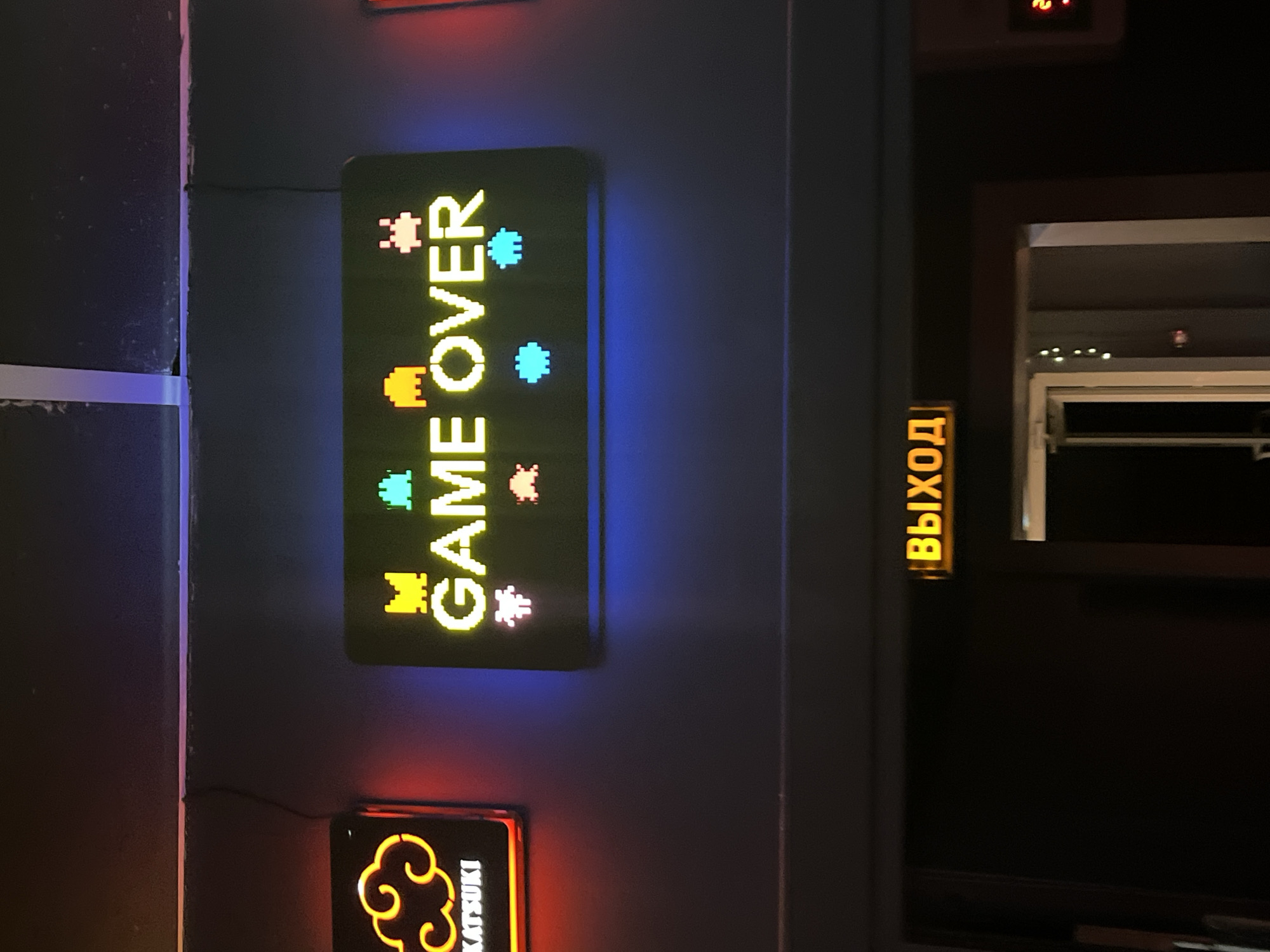 The image shows a vertical "GAME OVER" sign in pixelated style on a dark wall. Above it is a rectangular sign and below is another sign with a cloud design that says "KATSUKI". To the right, there is a vertical illuminated sign in Russian that says "EXIT." The background shows a dimly lit interior space, possibly a building in Chaikovsky, Russia, based on the Russian text on the exit sign. A partially visible window or opening is located to the right.