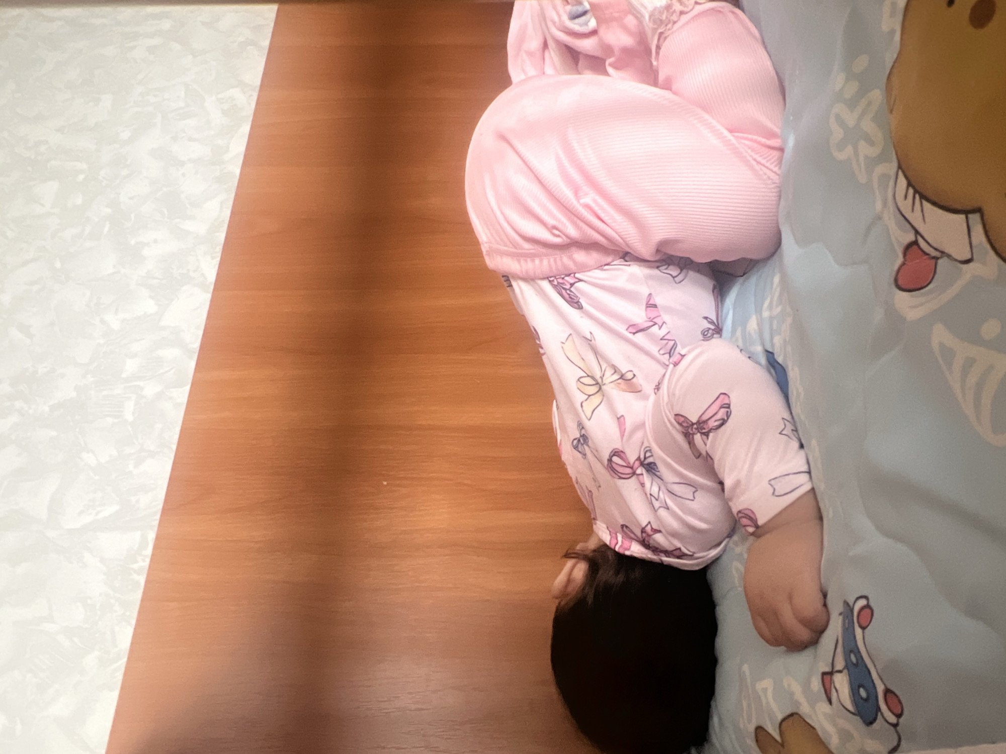 A child in pink pajamas is asleep on a bed, upside down, with their head near the floor and feet on a pillow. The background includes a wood floor and a light-colored wall. The location is not identifiable as Elena, Bulgaria, from the image alone.