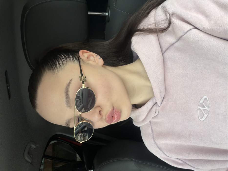 A female individual is depicted from the upper torso upward, seated within the interior of a vehicle, in what is identified by prompt as the city of Pernik, Bulgaria. She has dark hair styled in a slicked-back ponytail, with visible baby hairs along the hairline. Her eyebrows are dark and shaped. She is wearing gold-framed, oval-shaped sunglasses with dark lenses; the temples of the sunglasses feature a distinct "Miu Miu" logo. Two small, silver-colored stud earrings are visible in her right earlobe. Her lips are pursed in a "kissing" pose. She is wearing a light pink or faded lavender hooded sweatshirt with an embroidered white logo, consisting of an abstract "WD" or similar design within an oval, centered on the chest. The vehicle's interior features dark-colored headrests, suggesting leather or synthetic upholstery. Reflections in the sunglasses lenses indicate an exterior environment, likely a street or urban setting.