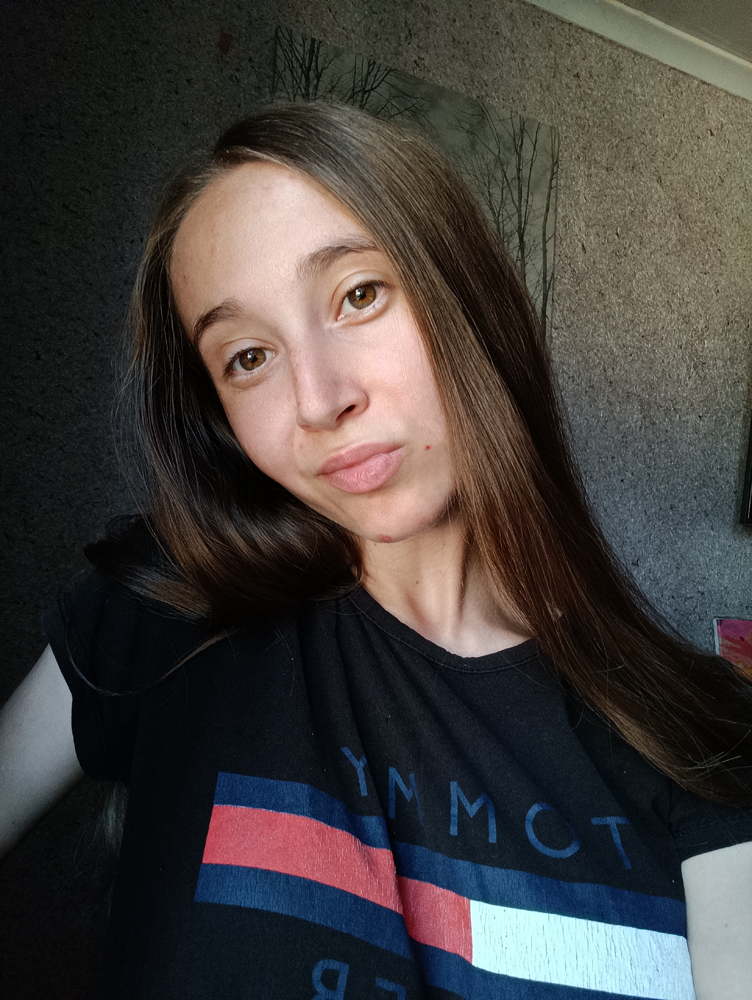 A young woman with long, brown hair and brown eyes is posing for a selfie. She is wearing a black t-shirt with a Tommy Hilfiger logo. The background is a textured, gray wall. The image appears to be taken indoors. No specific location in Vypasne, Ukraine is identifiable from the image.