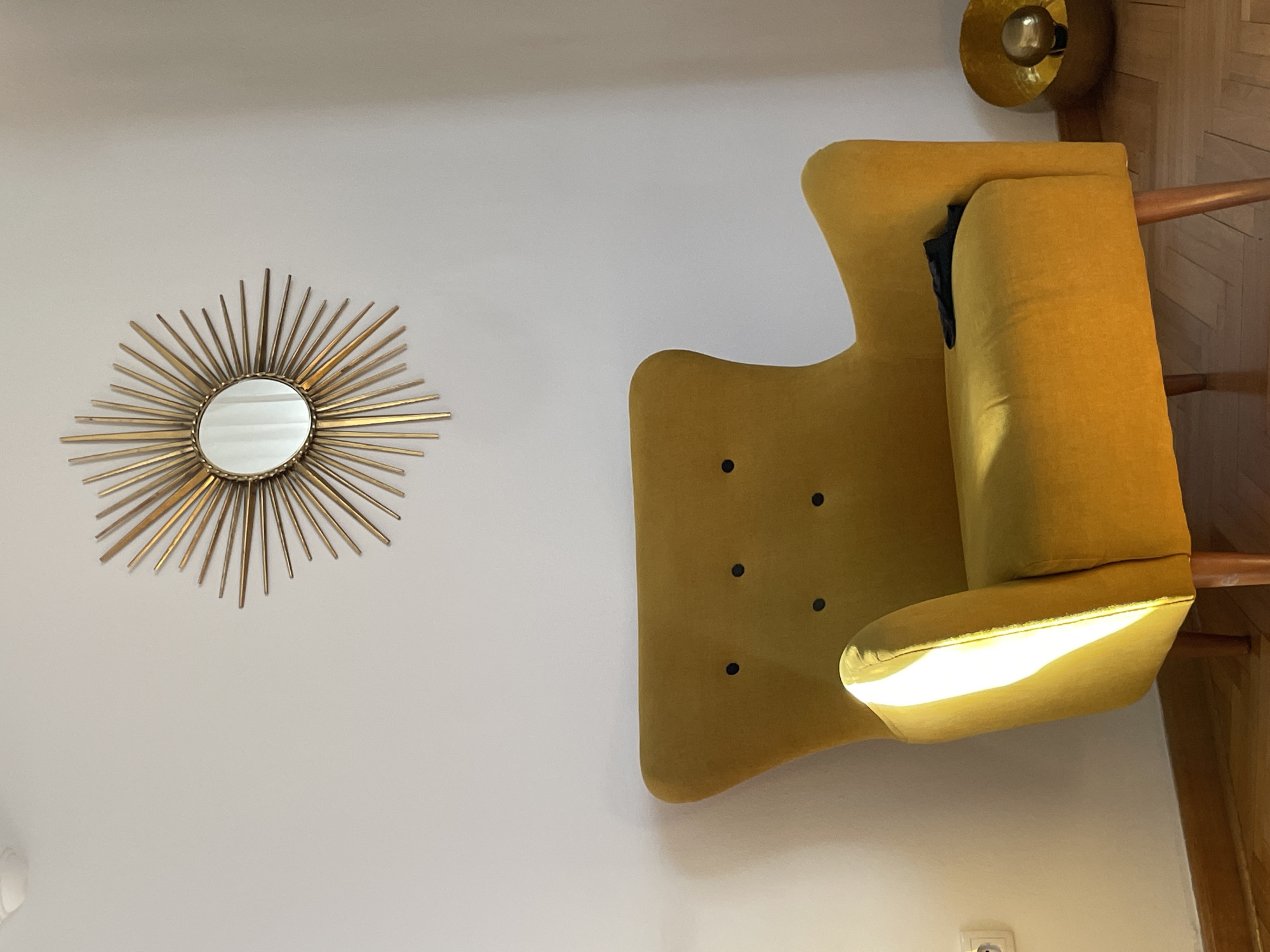 A sunburst mirror hangs on a white wall above a mustard yellow armchair. The armchair has black tufting on the back and a black object, possibly a piece of clothing, is placed on the seat. To the right of the armchair, a gold metallic bowl-shaped lamp with a gold bulb is visible. The floor is made of light-colored wood planks. Sunlight streams in from the left, illuminating the side of the armchair and casting a shadow on the wall. A white electrical outlet is visible on the wall to the left of the armchair. The scene appears to be an interior shot of a room.