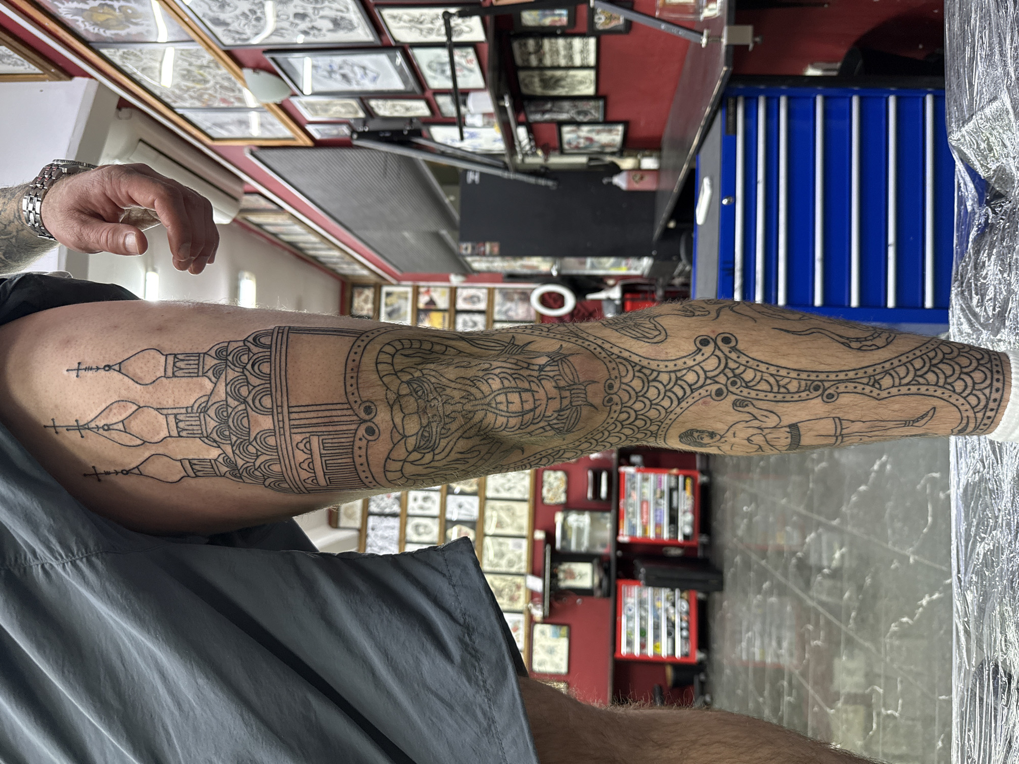 A man's tattooed leg is prominently displayed in the foreground. The tattoos are intricate black line drawings depicting a Russian Orthodox church with domes and crosses near the upper thigh, moving down to include what appears to be a large sea creature with a boat on its back and a boxer lying down. The man is wearing gray shorts and has visible arm hair. The background appears to be inside a tattoo parlor or studio. The walls are lined with framed tattoo designs, and there's a red color scheme in parts of the studio. A blue tool chest and some other equipment are also visible. The image is taken from a low angle, looking upward. The tattoos suggest a possible cultural or personal connection to Russia.