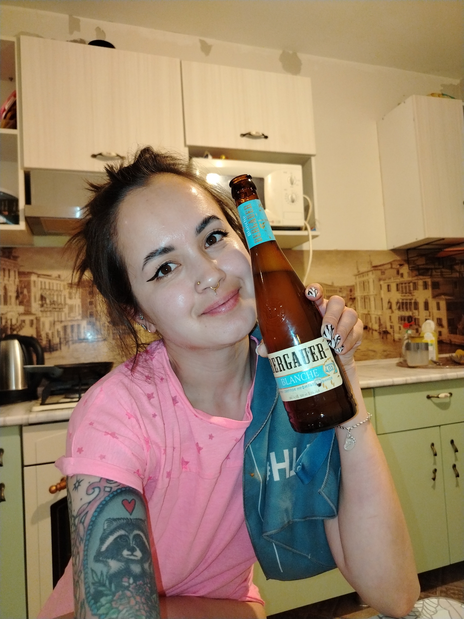A woman is in a kitchen, holding a brown beer bottle with a blue label. She has brown hair pulled back, brown eyes with winged eyeliner, and a nose ring. She is wearing a pink t-shirt. Her left arm has a tattoo of a raccoon. Her right hand, which holds the bottle, has black and white patterned nails. A blue towel is draped over her arm. The beer label is in Russian.
The kitchen has light green cabinets with dark handles and a countertop with a picture of a Venetian cityscape. Behind her is a microwave and beige cabinets. A silver tea kettle is visible on the counter. The kitchen could be located in the city of Noviy, Russia.