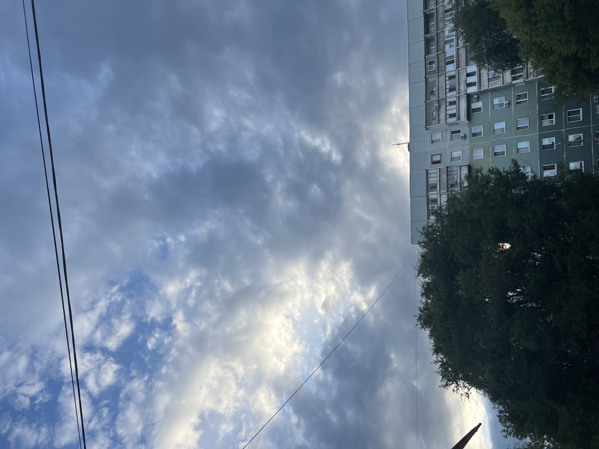 The image shows a tall, multi-story apartment building in Ulyanovsk, Russia, under a cloudy sky. The building has a light teal facade with numerous windows and balconies. Dense green trees are visible in the foreground and on the right side of the building, partially obscuring it. Two thin black wires stretch horizontally across the top of the frame, and a single wire diagonally crosses the sky. The lighting suggests it is either early morning or late evening, with the sun partially breaking through the clouds in the upper left quadrant of the sky. No people are visible in the image.