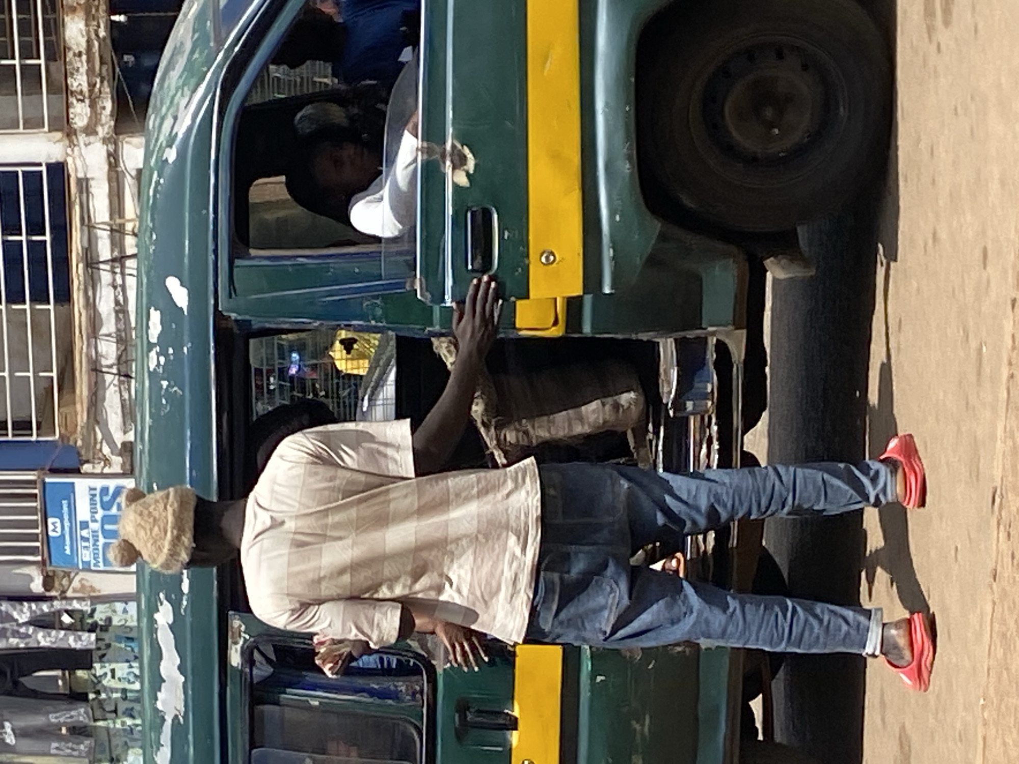 In Bukuru, Nigeria, a man wearing a light brown striped t-shirt, blue jeans, and red clogs stands beside the open passenger door of a dark green van with a yellow stripe. He is wearing a tan beanie with two pom-poms. His left hand rests on the van's door frame, and his right hand is visible, partially obscured by his body. Inside the van, a person in a white shirt is looking towards the right, and another person in a blue shirt is seated behind them. The van's paint is chipped in several places, and there is a large brown sack behind the open door. In the background, a building with white bars across its windows and a blue sign with white lettering is visible.