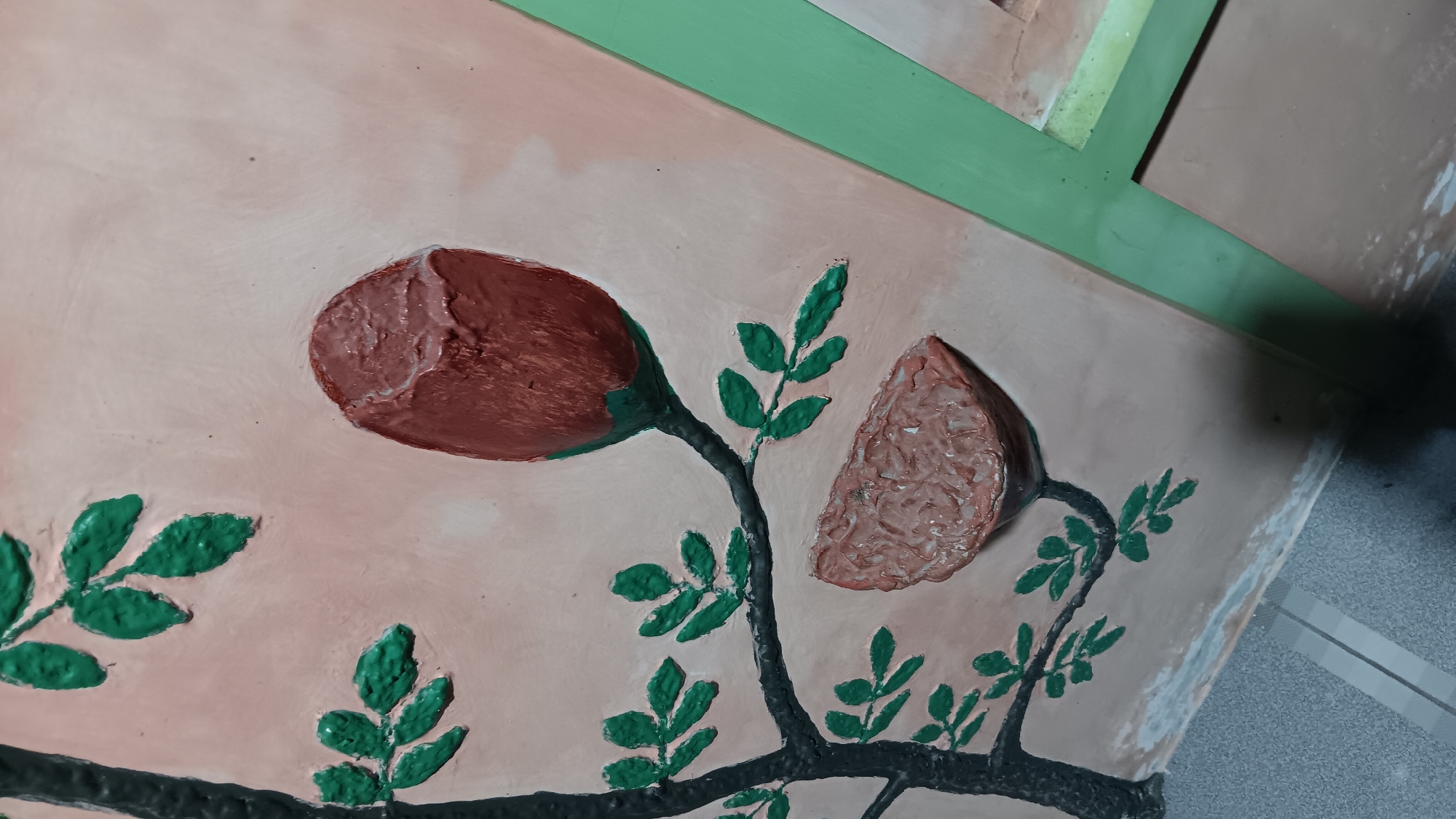 The image is a close-up of a decorative relief on a wall. The relief depicts a stylized tree branch with green leaves, a red rosebud, and a detached petal or fruit. The background of the relief is a pale peach color. To the right of the relief, a section of a light green frame is visible. The lower portion of the image shows the texture of the floor, which appears to be tiled with grey and white rectangular tiles. The overall impression is of an artistic embellishment on a wall, likely in a public or semi-public space. The location in Sonobekel, Indonesia, suggests it is an outdoor or semi-outdoor mural.
