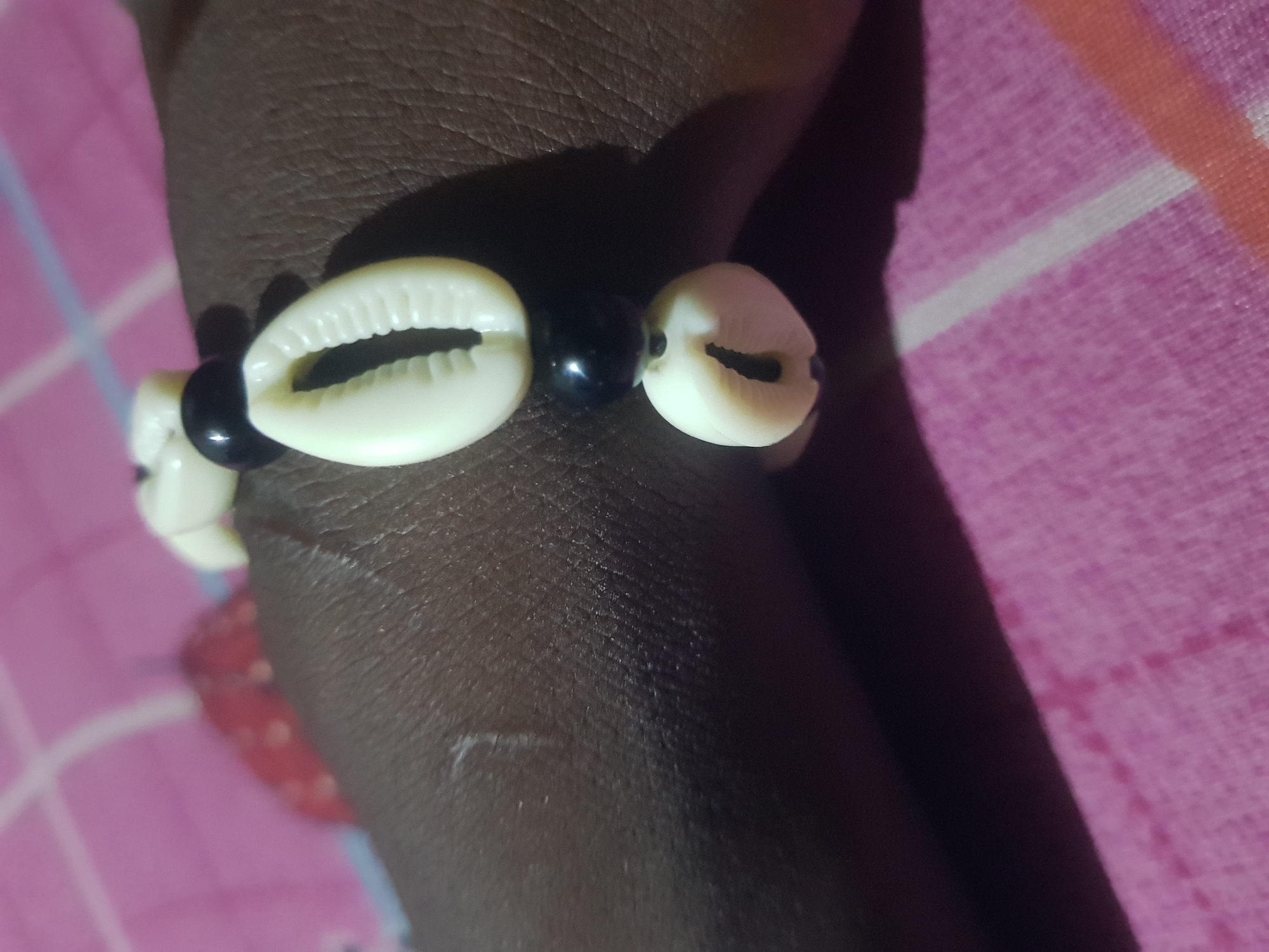 A bracelet made of white cowrie shells and black beads is worn on a dark-skinned wrist. The bracelet is positioned diagonally across the wrist, with the shells and beads clearly visible. The background consists of a pink fabric with white and orange stripes. The lighting creates shadows on the skin and the bracelet. The scene appears to be indoors, possibly on a bed or sofa.