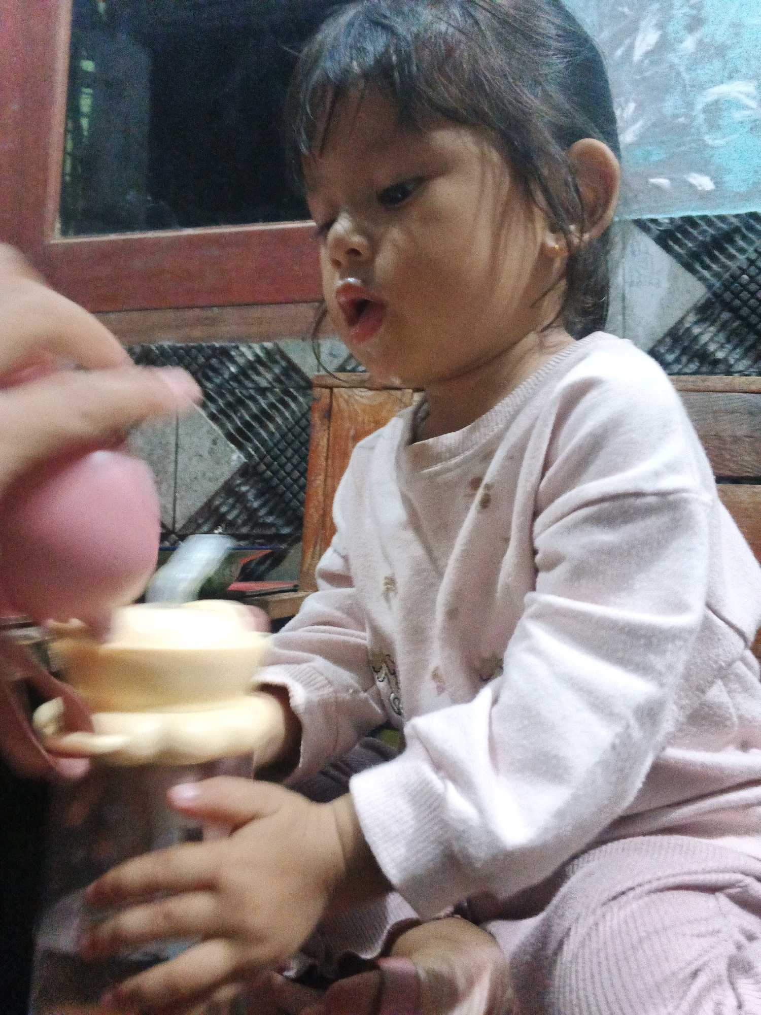 An infant, seated and wearing a pale pink long-sleeved top with a delicate embroidered pattern, is positioned in the foreground. The infant's mouth is open, suggesting an action or vocalization. To the left of the infant, a hand, partially visible, holds a pink object and interacts with a light yellow toy, which appears to be a stacking ring toy. The background features a dark, reflective surface, possibly a window or mirror, and wooden furniture, suggesting an indoor residential setting. The overall scene is dimly lit, with a slight graininess to the image. The context suggests a domestic activity, likely play or feeding, in an unspecified residential location within the city of Kadu, Indonesia.