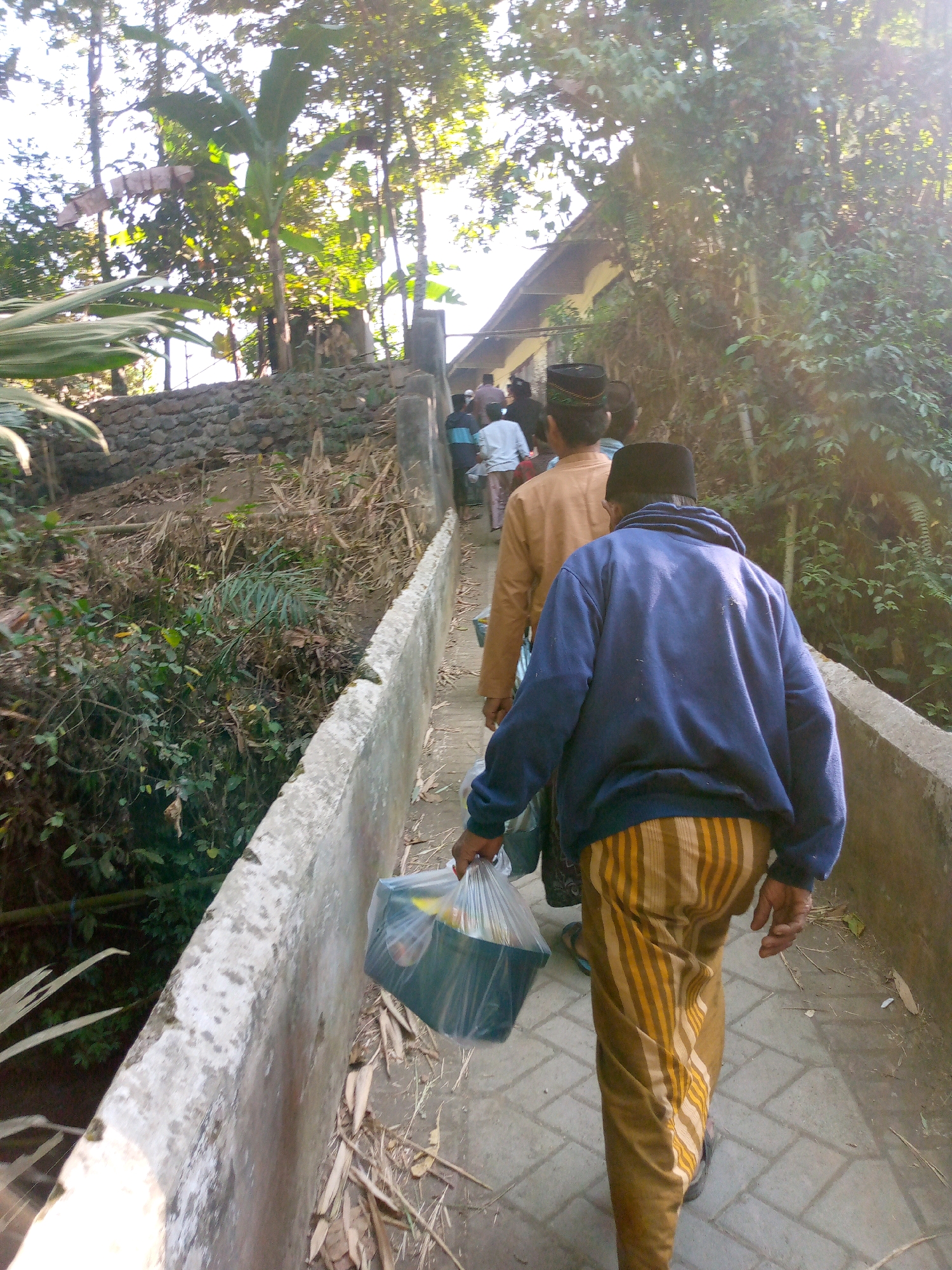The image depicts a group of people walking along a paved pathway bordered by a low concrete wall. The pathway appears to be elevated, with vegetation and trees on either side. The overall environment suggests a rural or semi-rural setting. The pathway is made of interlocking bricks.
In the foreground, a man is walking away from the camera, wearing a blue hooded sweatshirt and a sarong with vertical stripes in shades of yellow and brown. He is carrying a plastic bag containing items. Behind him, another man in a tan shirt and black cap is also walking away from the camera. Further ahead, several other individuals are visible, some wearing caps, walking up the pathway towards a building with a sloping roof, partially obscured by trees and foliage. The building appears to be a simple structure with a dark roof.
The vegetation is dense, with large green leaves and tall trees, suggesting a tropical or subtropical climate. Sunlight filters through the leaves, creating highlights and shadows, indicating it is daytime. The presence of a stone wall, possibly part of a retaining structure, is noted on the left side of the pathway. The scene is consistent with the location of Jambe Arum, Indonesia, characterized by its lush greenery and common architectural styles for local buildings and pathways.