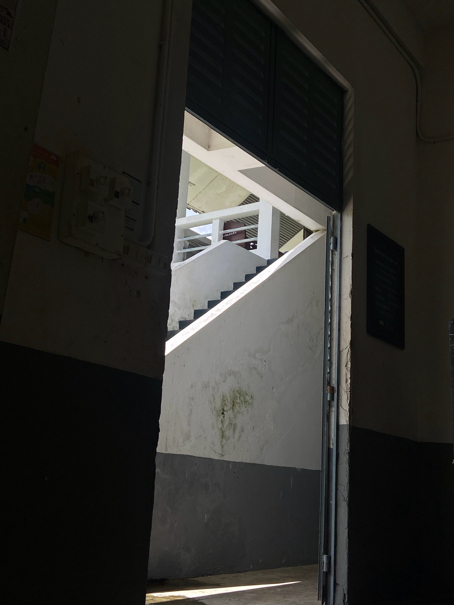 The image depicts an interior view from a doorway looking out towards a concrete staircase with a white railing. The doorway is on the left side of the frame, and its interior wall is painted dark gray at the bottom and a lighter color above. Electrical conduits and a light switch panel are visible on the left wall. The doorway opens into a hallway or corridor. The staircase is illuminated by natural light streaming in from above and to the right, creating strong shadows. The walls of the staircase area are white, with some staining visible. There are dark shutters or louvers visible at the top of the opening leading to the staircase. A dark rectangular plaque with text is mounted on the right-hand wall of the corridor. The overall impression is one of a utilitarian, possibly institutional building, with an emphasis on concrete construction and natural lighting. The location of Ban Si Yaek San Nare San, Thailand, is not explicitly visible or inferable from the image itself.